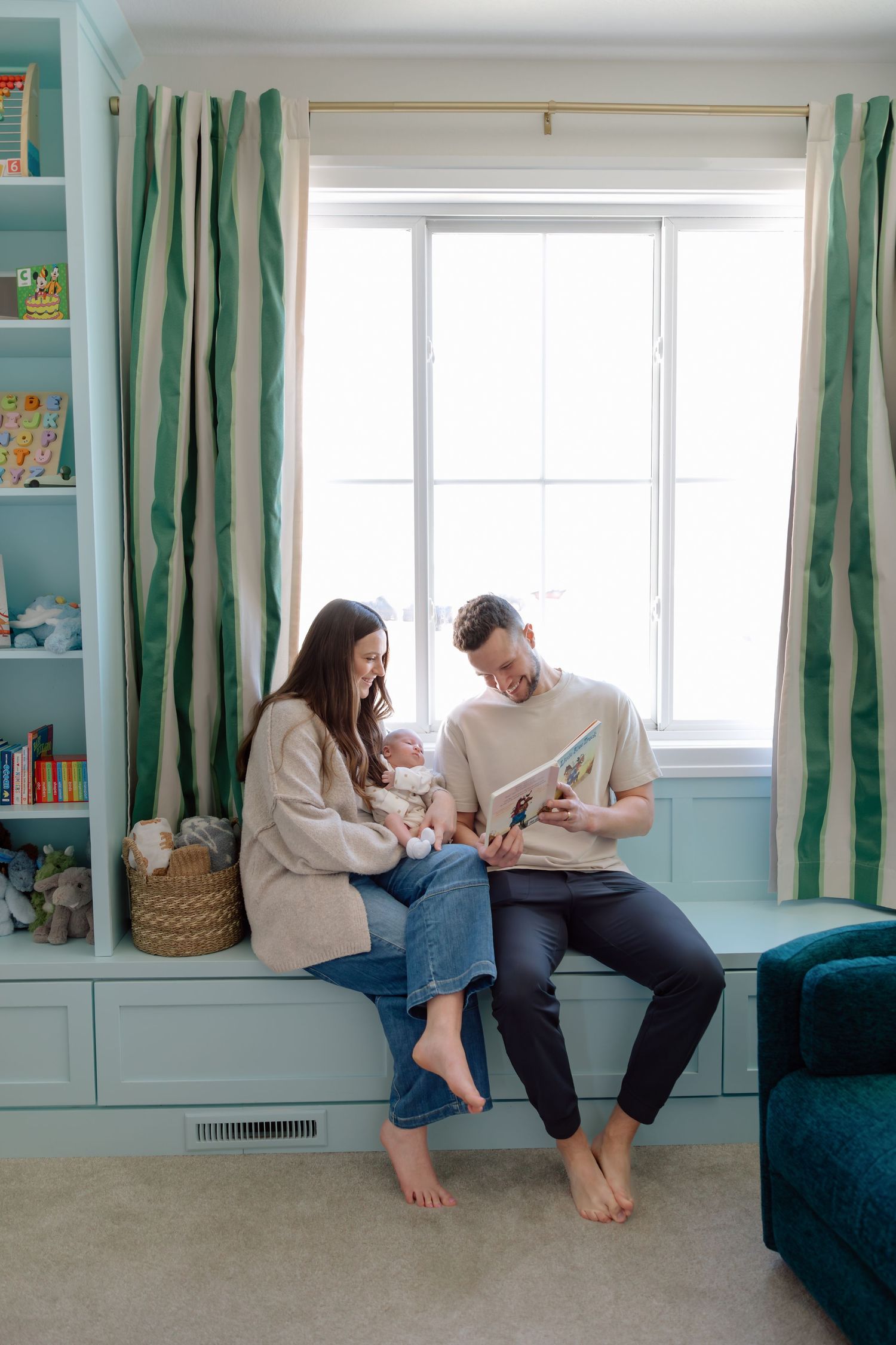 Young family reading a book together with newborn on a window seat in a nursery.