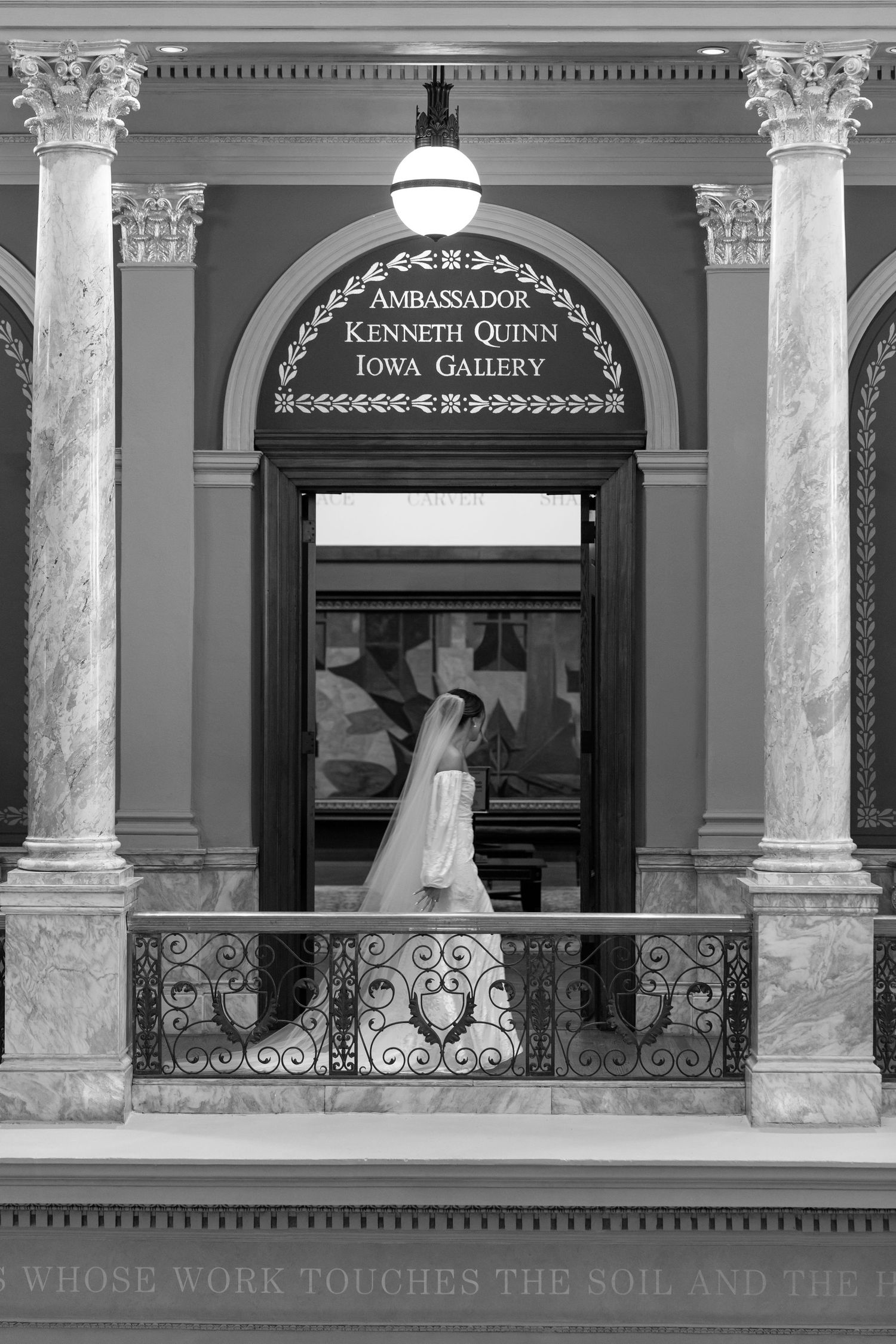 Bride in wedding dress and veil stands on ornate balcony of Ambassador Kenneth Quinn Iowa Gallery in black and white photo.