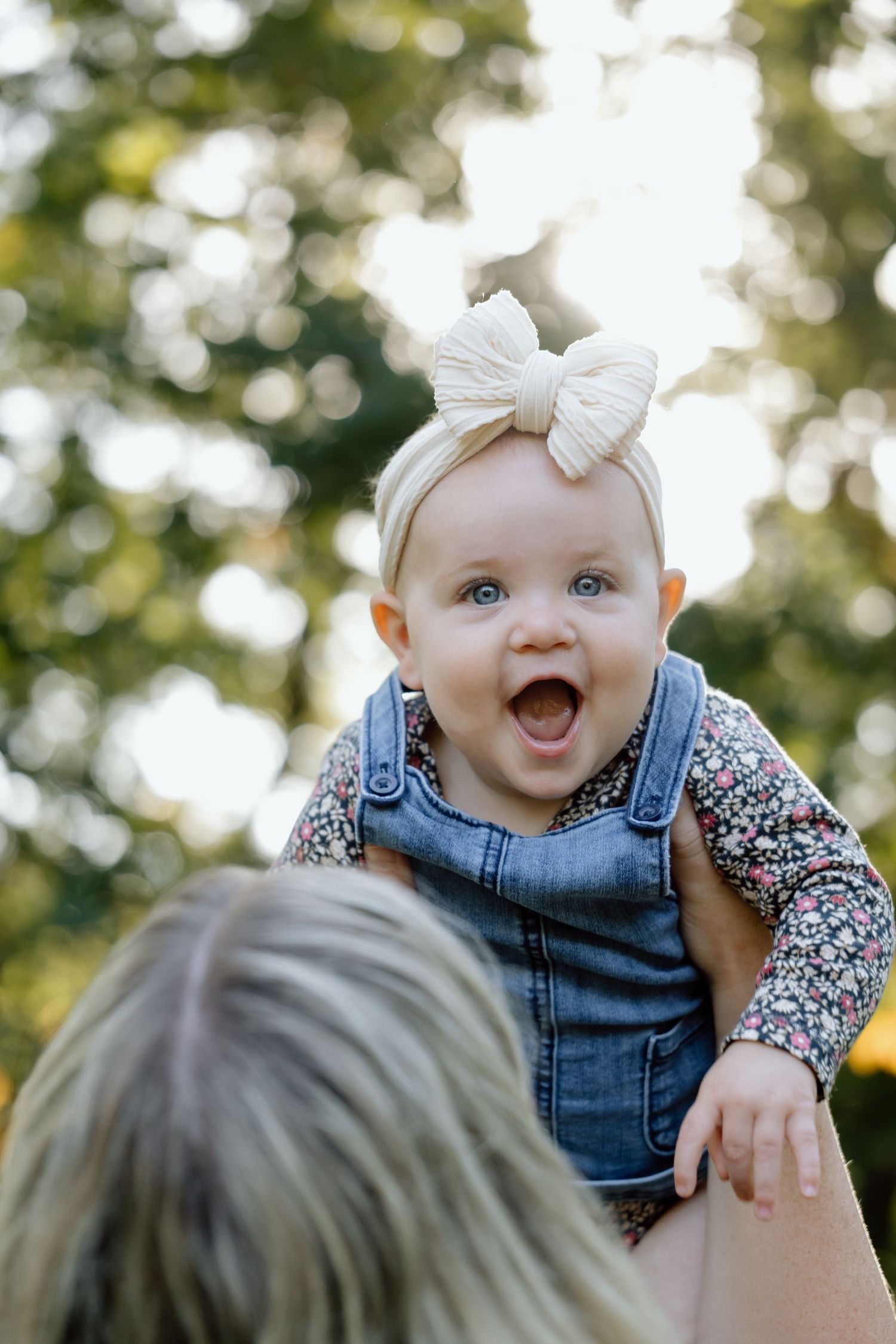 Joyful baby girl in denim overalls and bow headband laughing while being lifted outdoors.