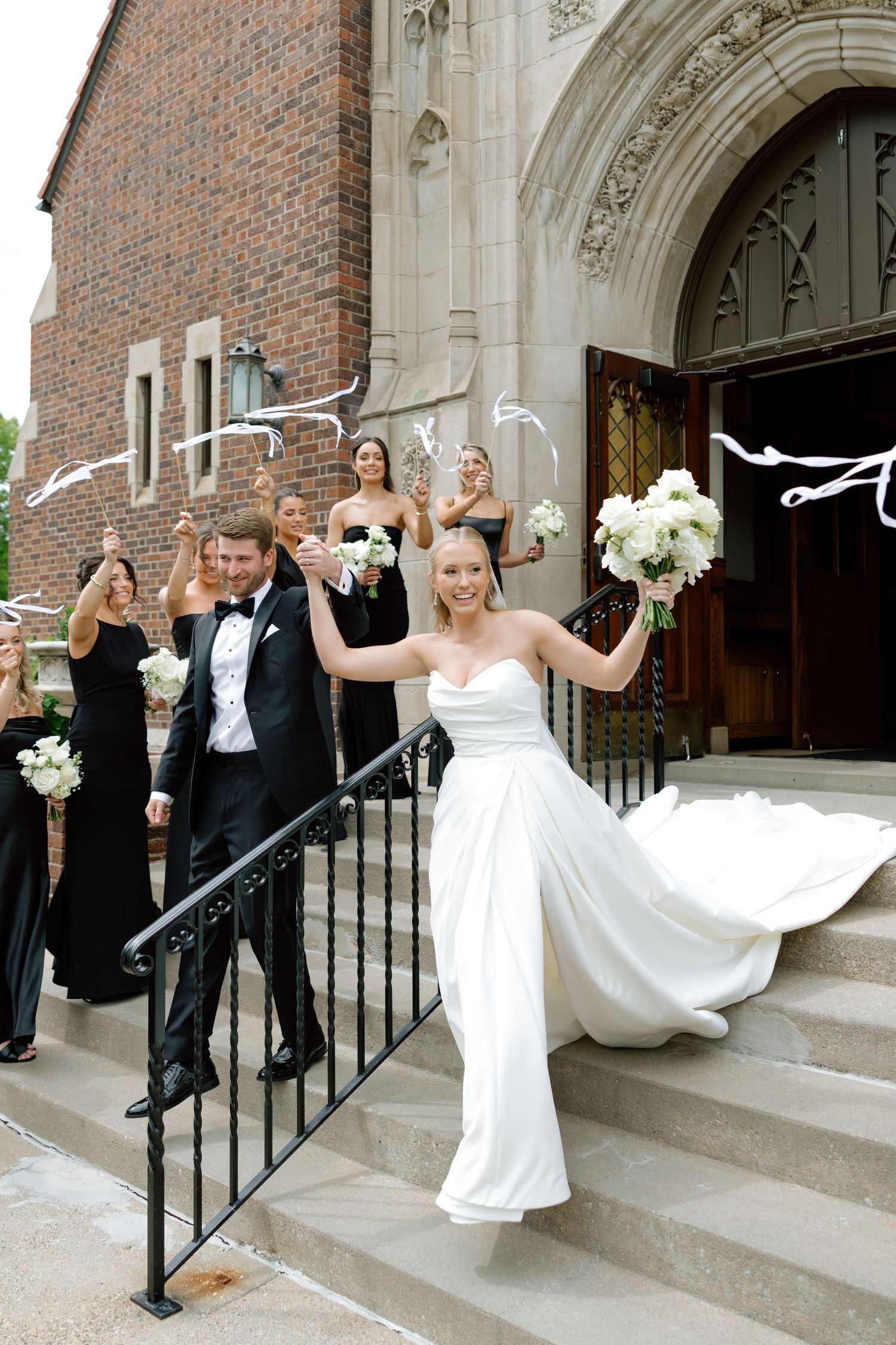 Bride and groom celebrate on church steps with wedding party in black attire holding white bouquets.