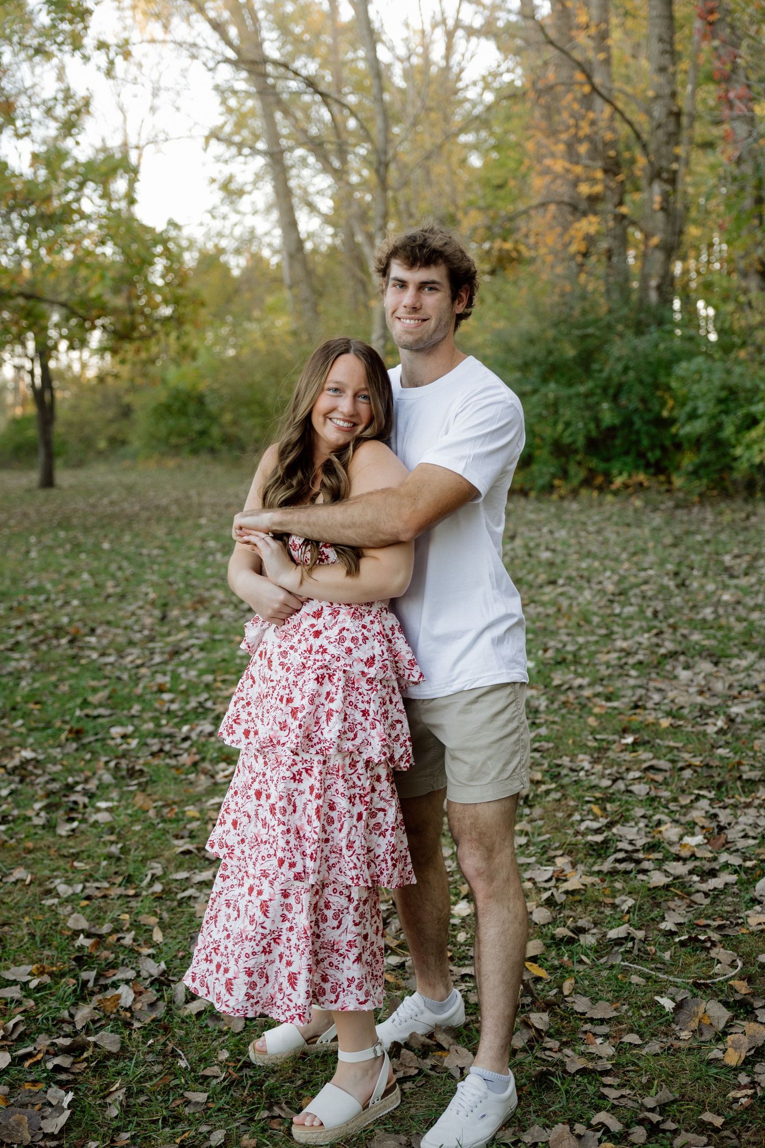 Happy couple embracing in a park surrounded by autumn leaves, woman in pink floral dress and man in white tee.