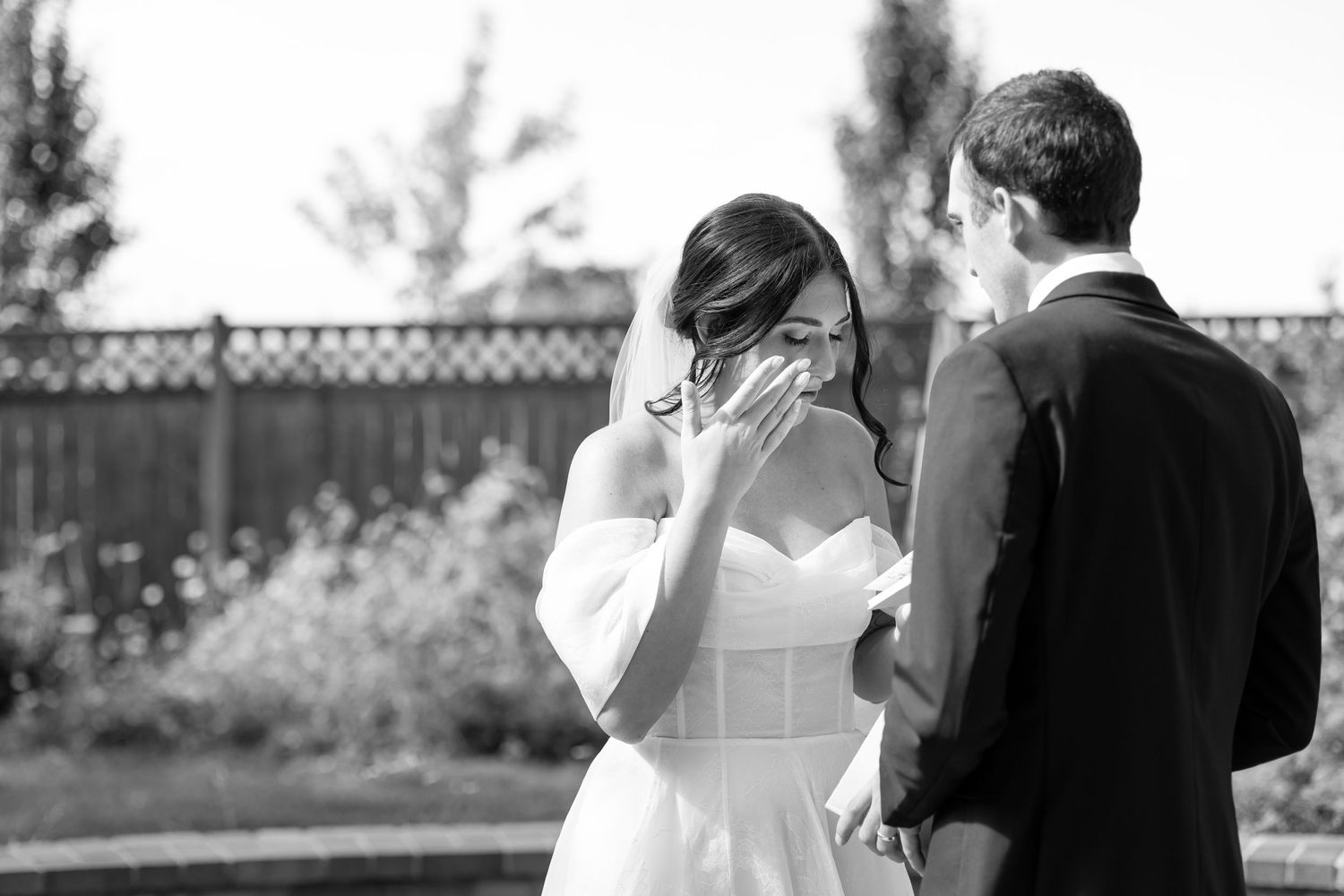 Emotional bride wiping tears during outdoor first look with groom in black suit at wedding.