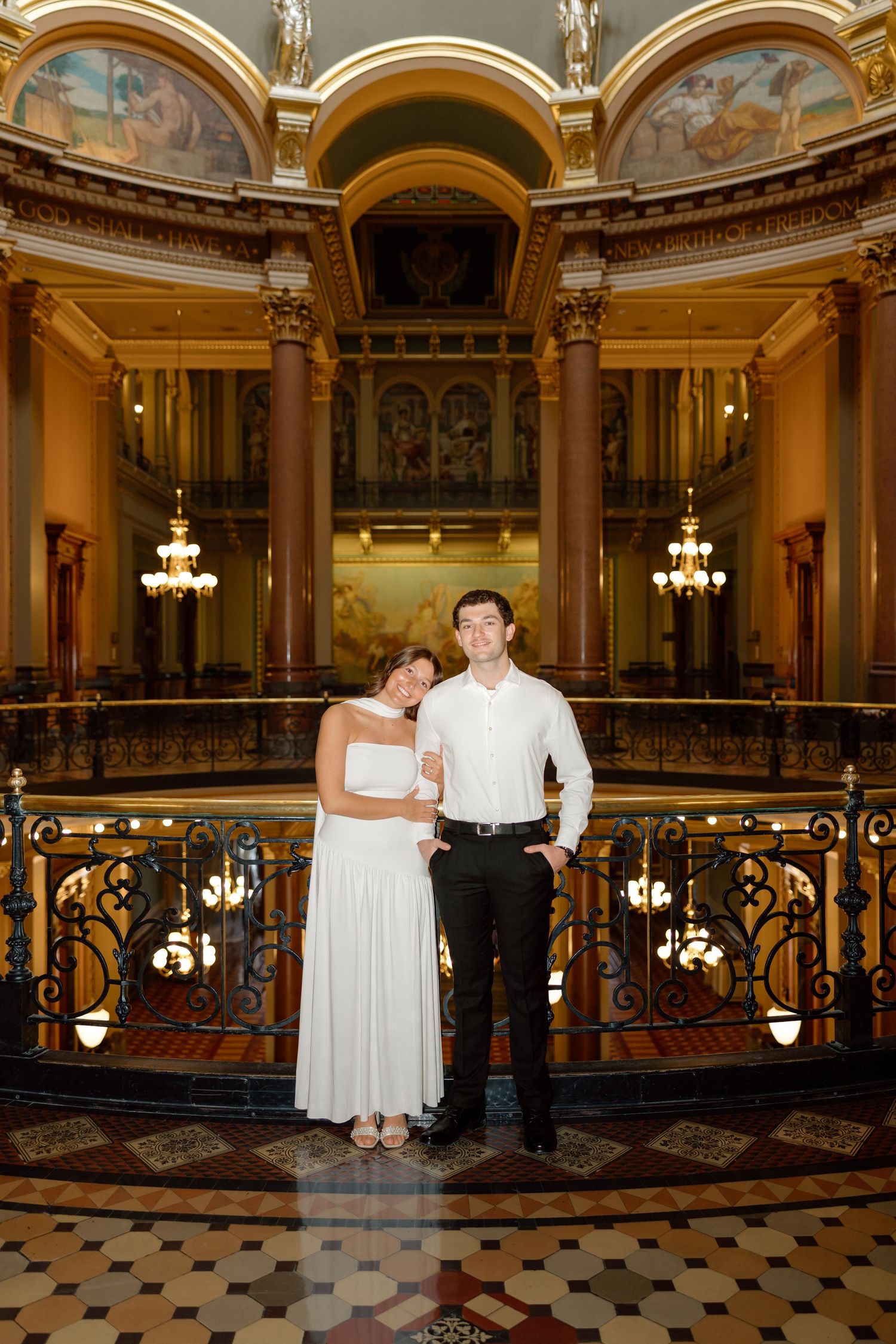 Elegant couple in white wedding attire poses inside a grand ornate rotunda with chandeliers.