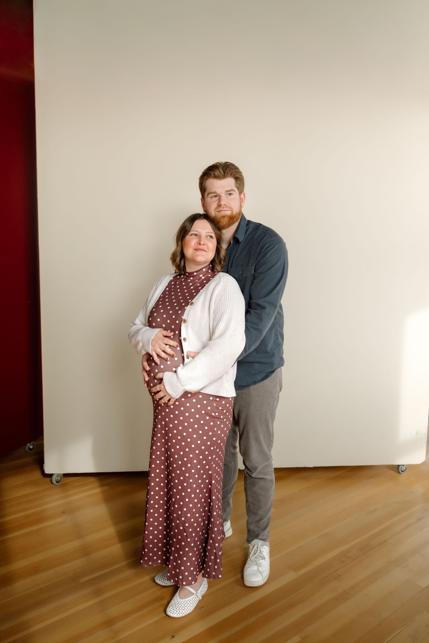 A couple poses together, the man embracing his pregnant partner in a polka dot dress against a white wall.