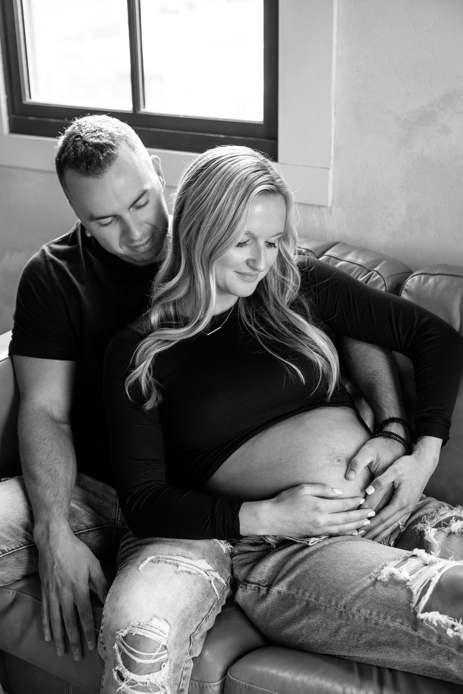 Black and white photo of a couple sitting on a couch with hands on pregnant belly.