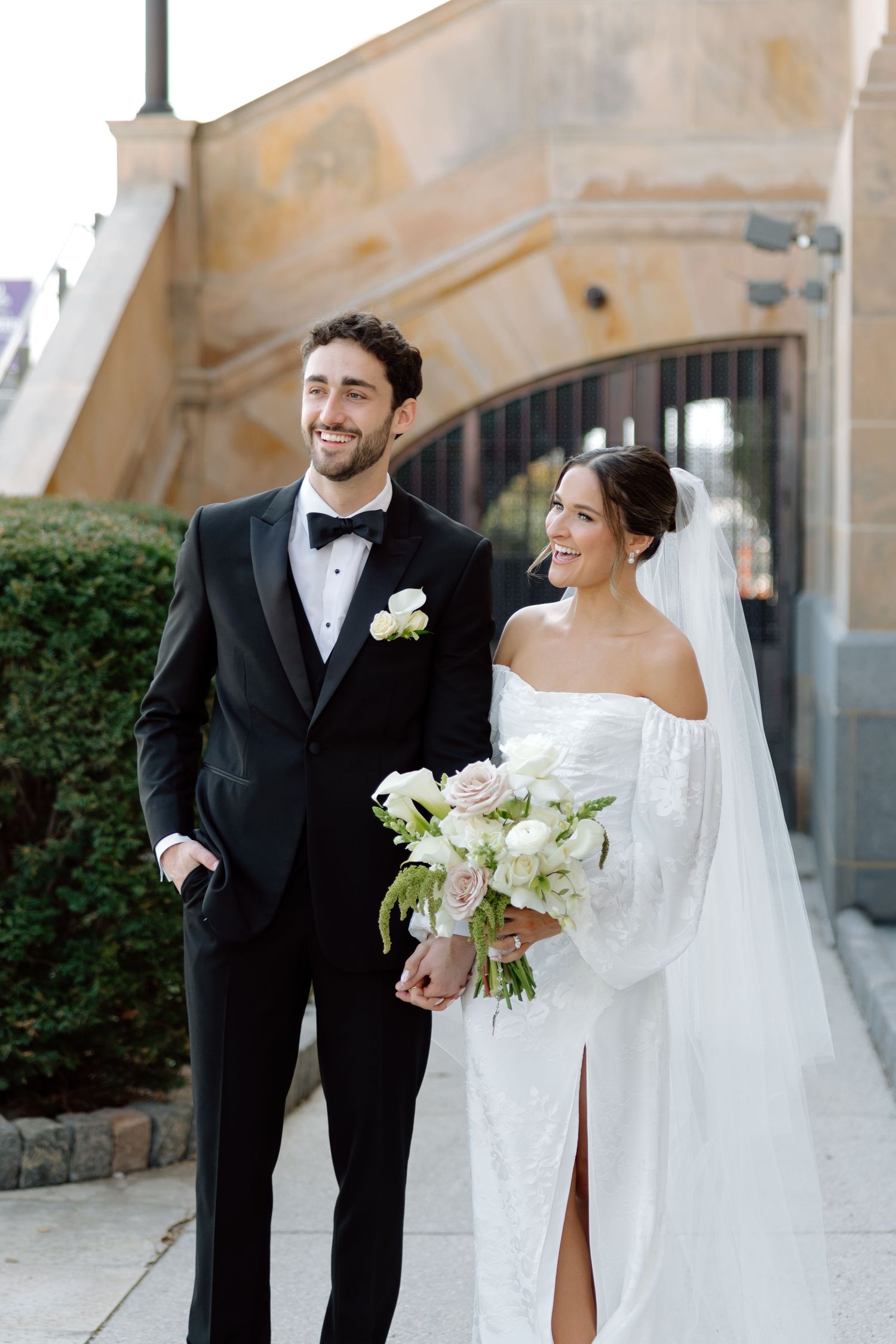Smiling bride and groom walking outdoors, bride holding white and pink bouquet in wedding attire.