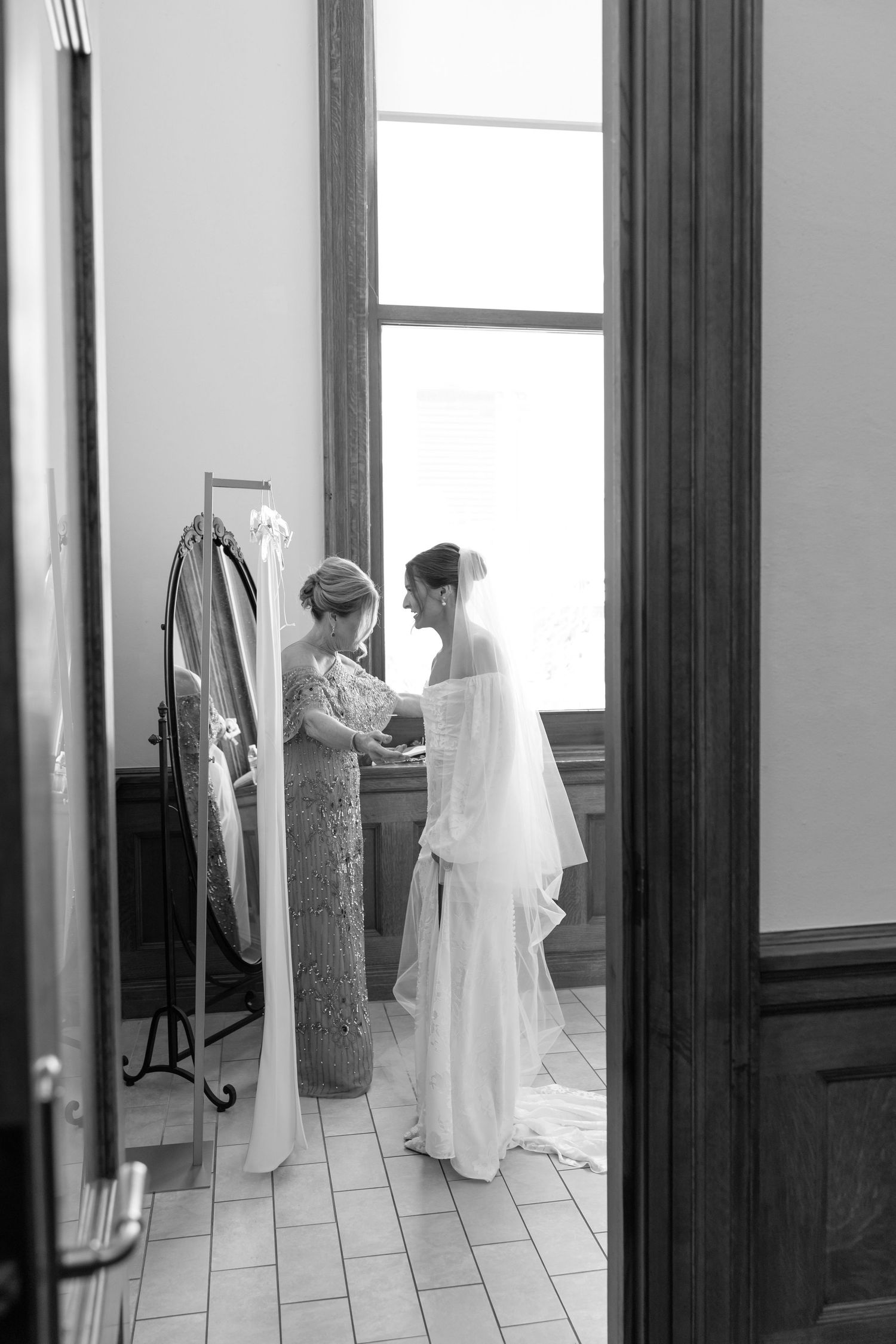 Bride in white gown and veil being helped by her mother near a bright window before the wedding.