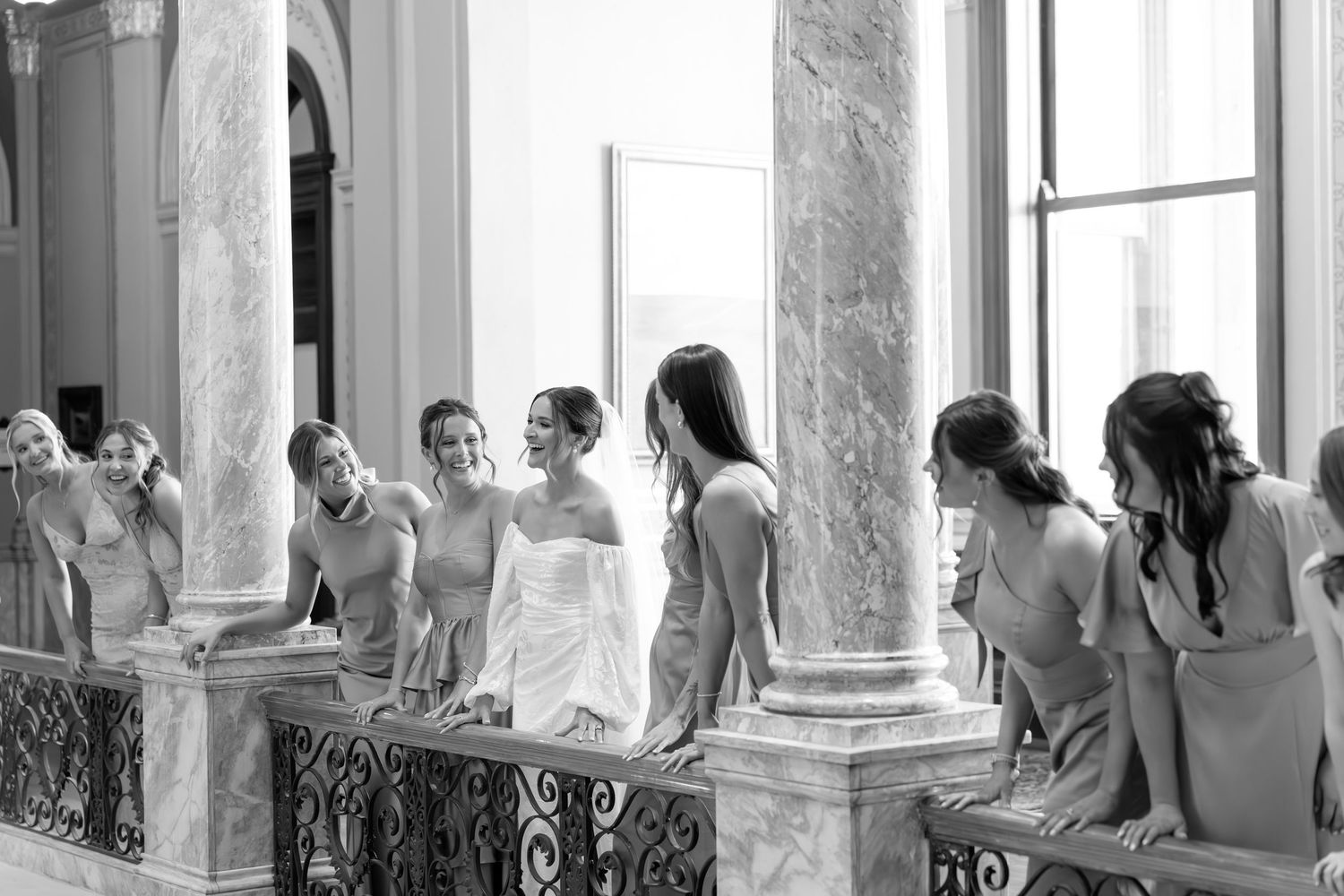 Black and white photo of bride with bridesmaids on elegant balcony with marble columns at wedding venue.