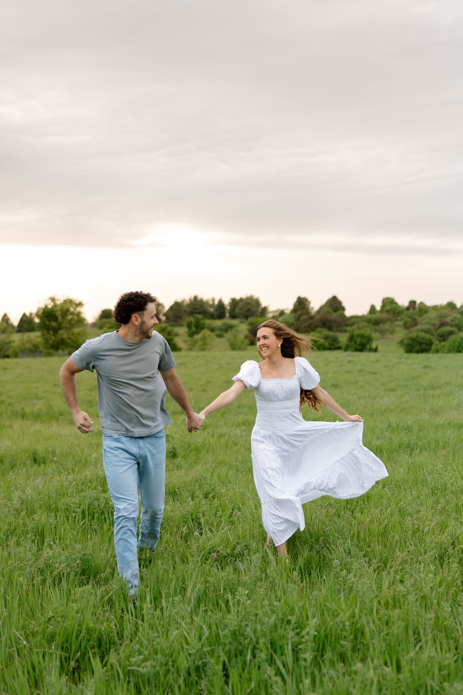A joyful couple runs hand-in-hand through a lush green field, the woman in a flowing white dress, smiling at each other.