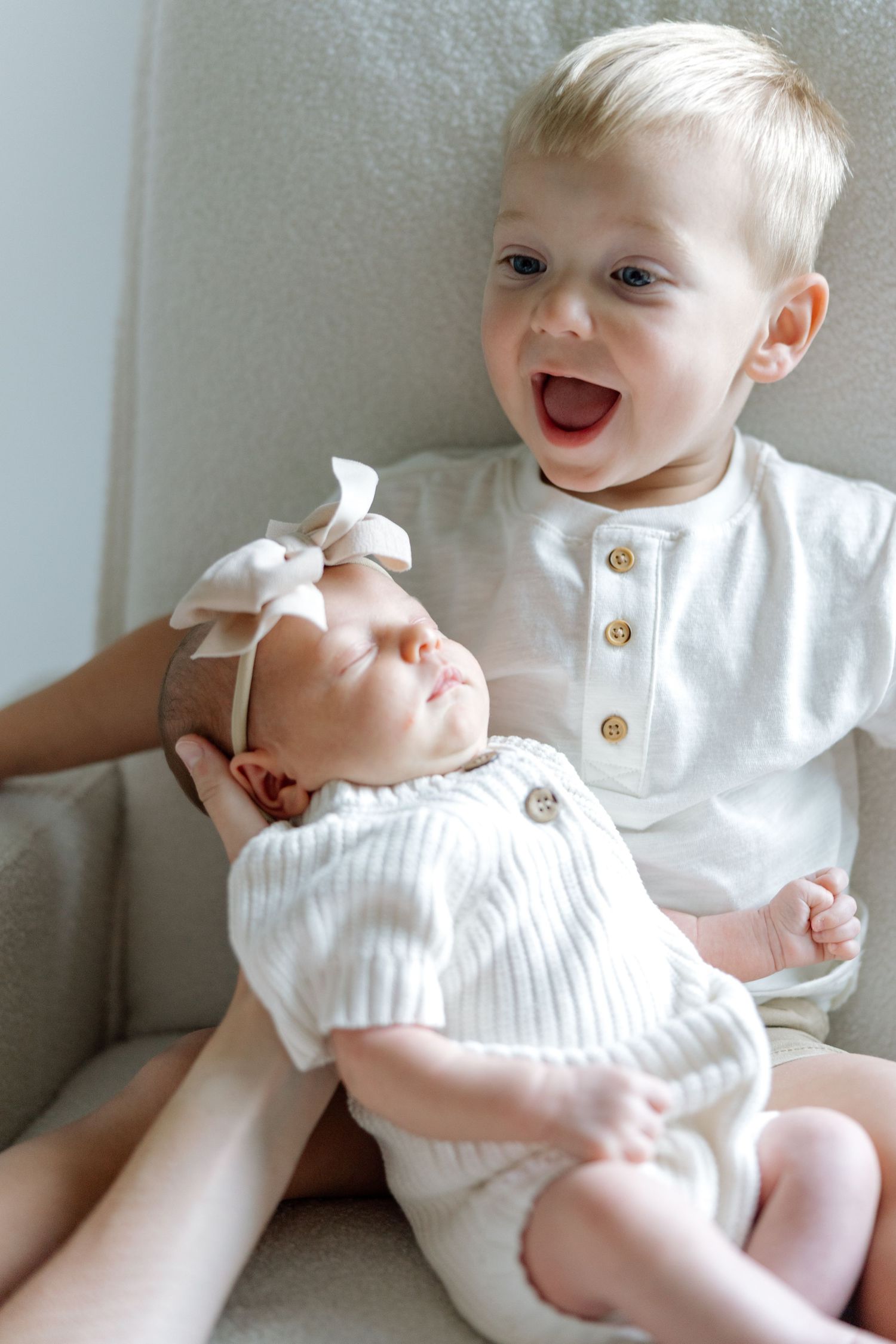 Excited toddler boy sitting with sleeping newborn baby sister in white outfits on chair.