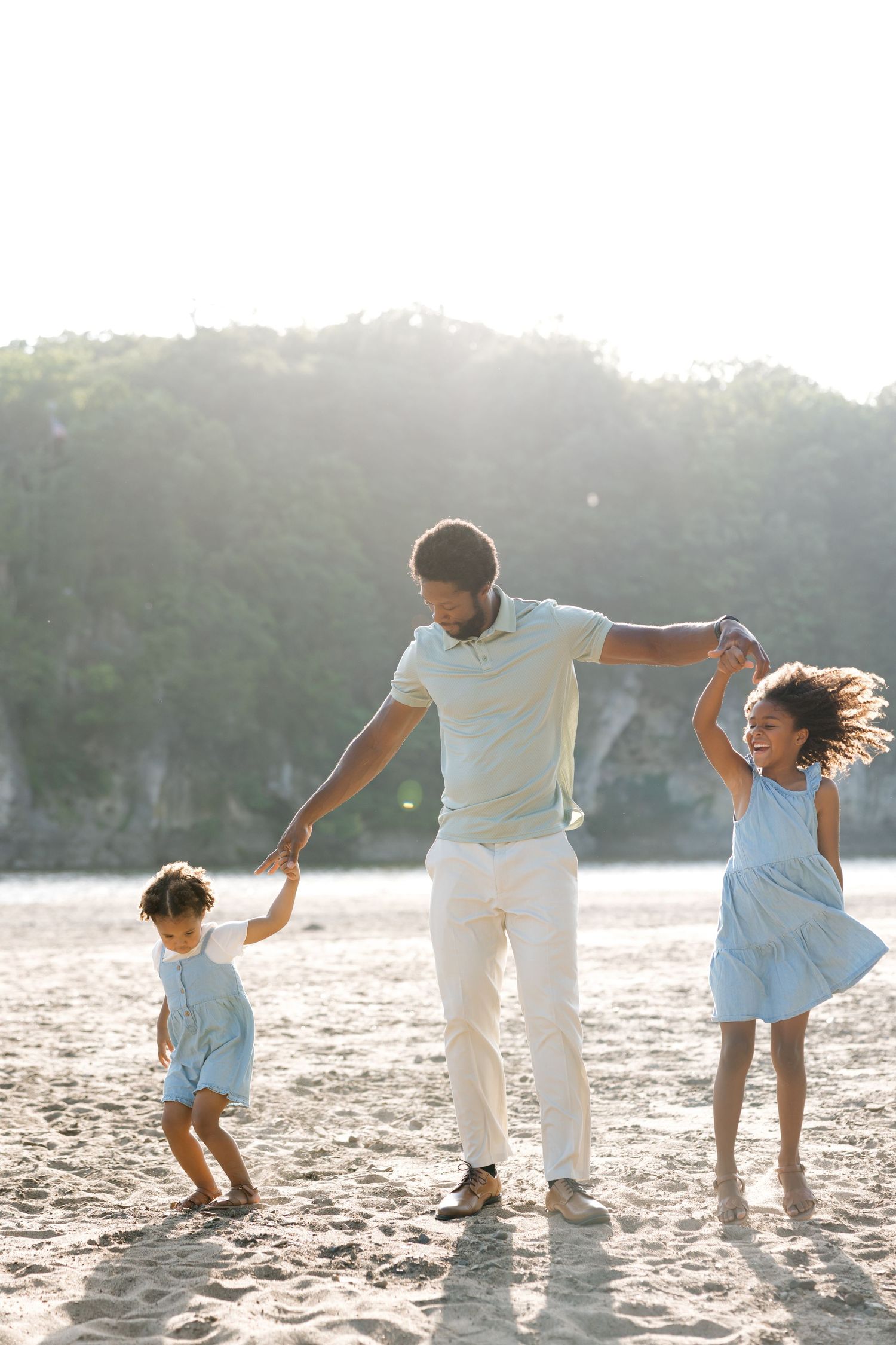 Father dancing on beach with two young children, holding hands in sunny coastal setting.