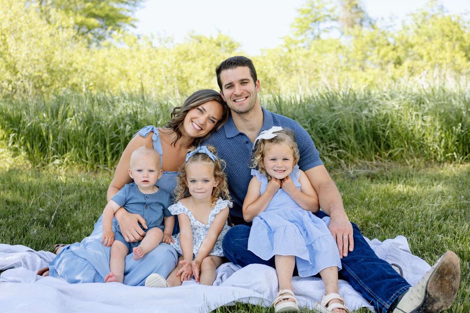 Family of five sitting on a blanket in blue outfits in a lush green outdoor setting.