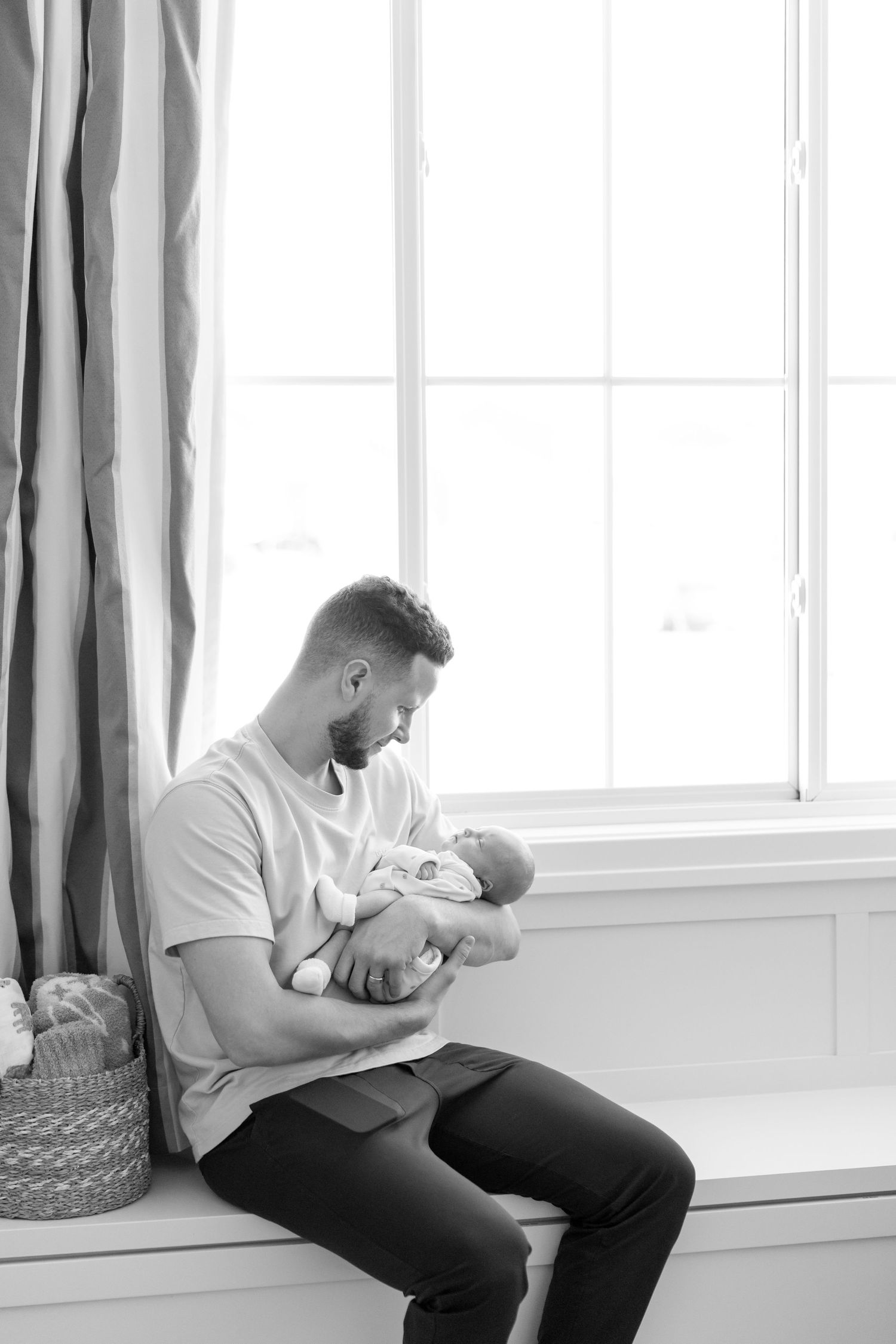 Black and white photo of a father sitting by a window tenderly holding his newborn.