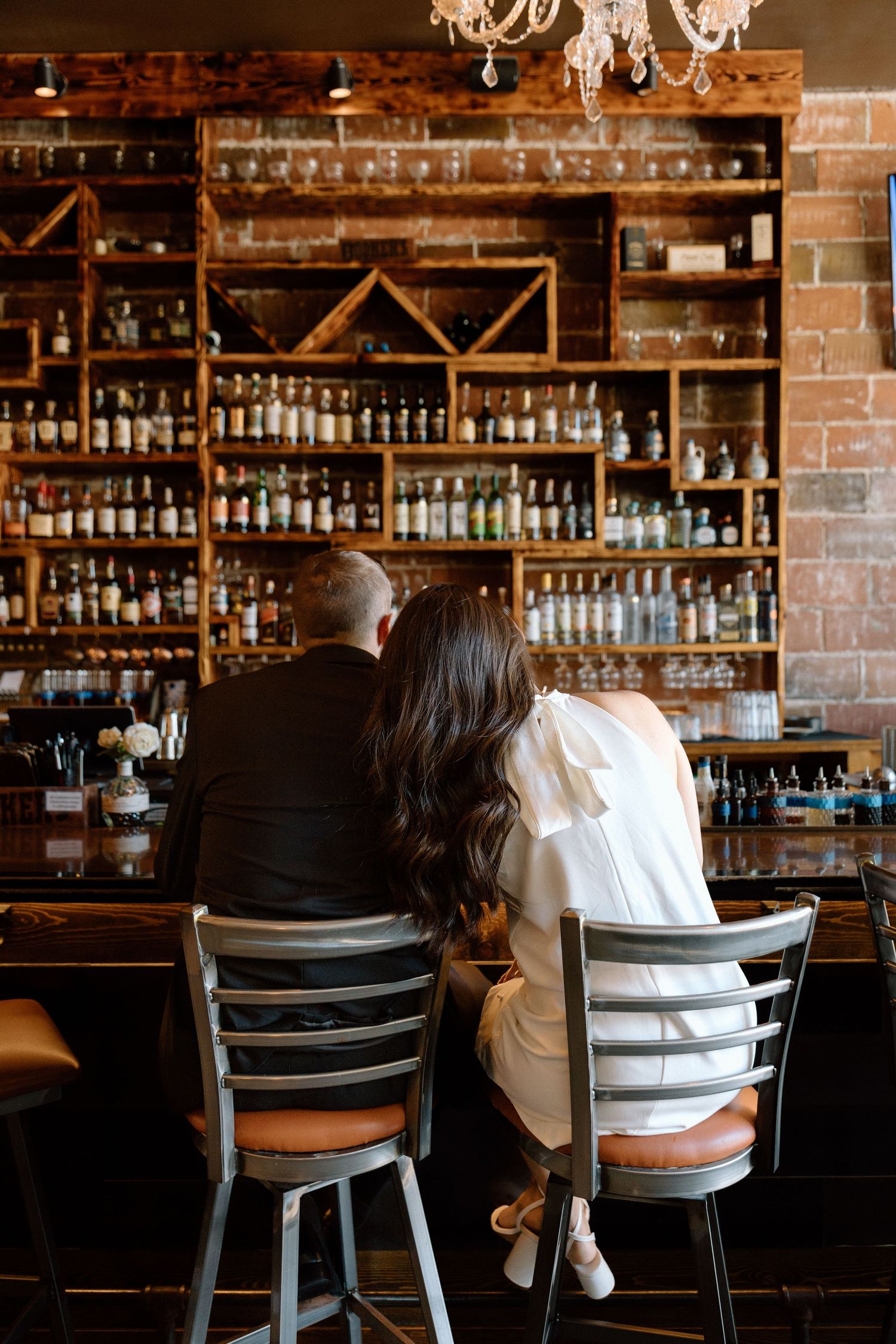 Couple sitting at a rustic bar with extensive bottle display, woman in white and man in black suit.