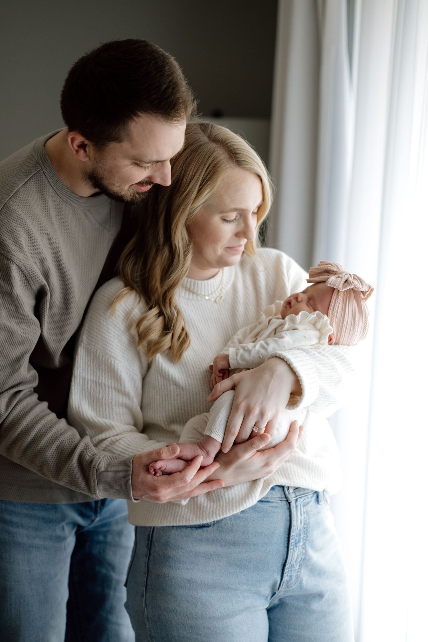 Happy new parents in jeans and sweaters lovingly gazing at newborn by window.