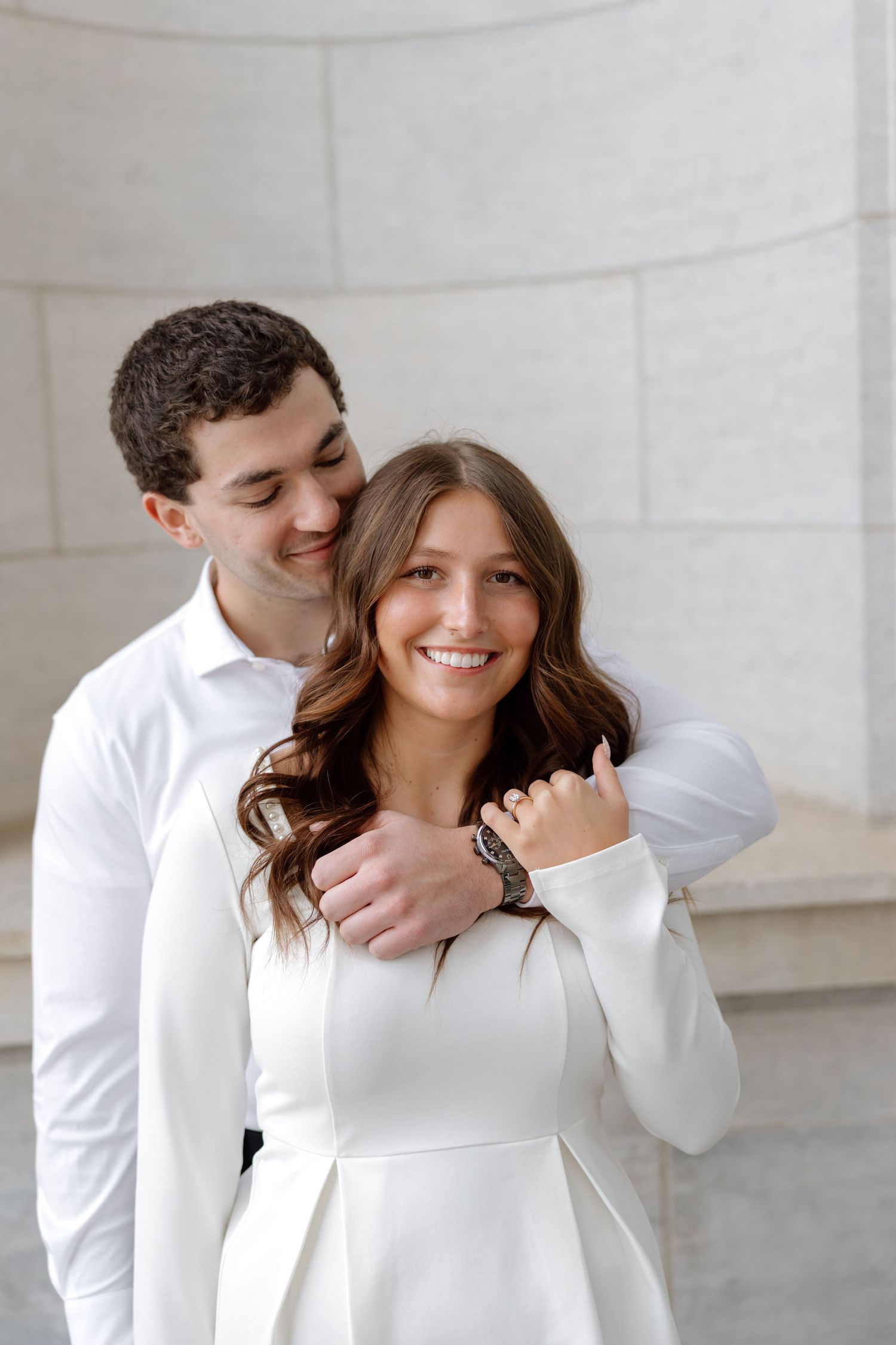 Man in white shirt embraces smiling woman in white dress from behind against a light stone column background.