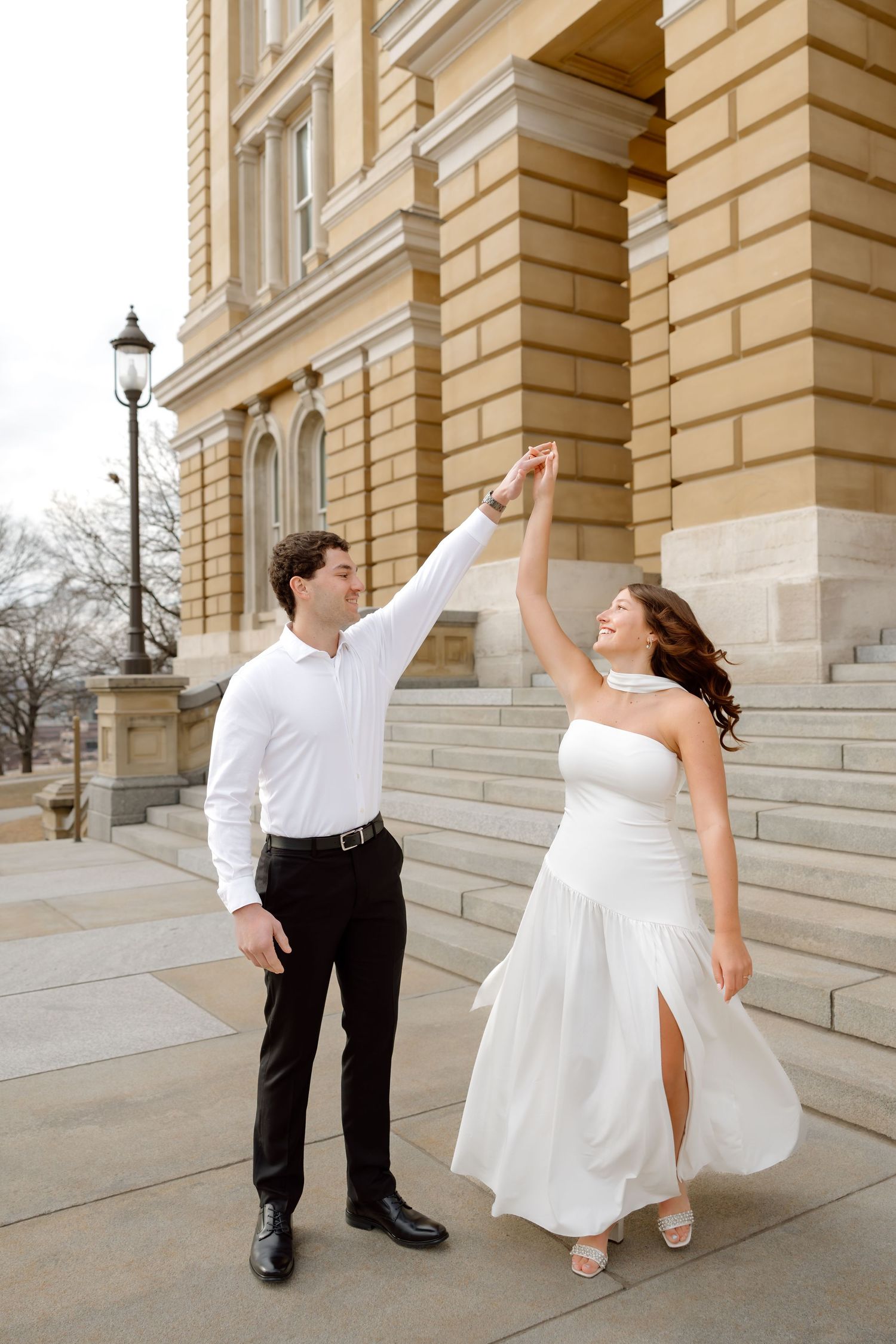 Couple dancing on steps of historic building, groom in white shirt spinning bride in elegant white slit dress.