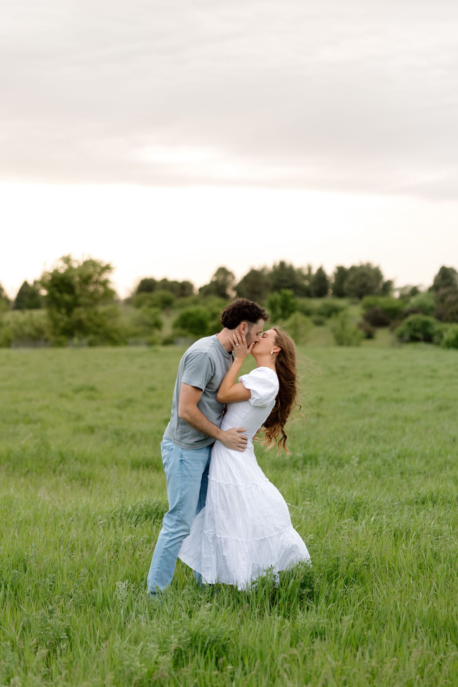 Couple sharing a romantic kiss in a lush green field, woman in white dress, man in jeans and gray shirt.