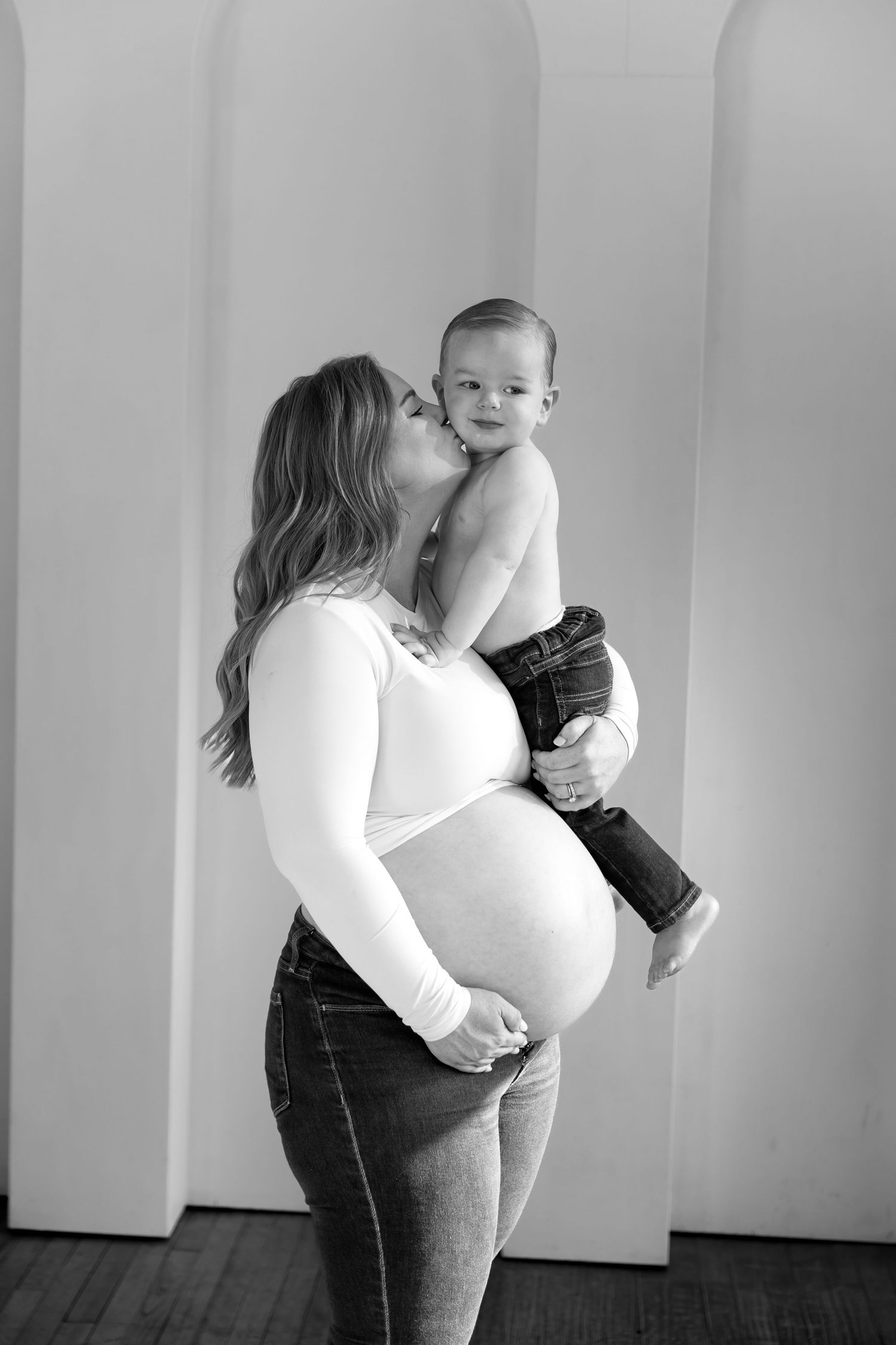 Black and white photo of a pregnant woman kissing her toddler son while cradling her baby bump.