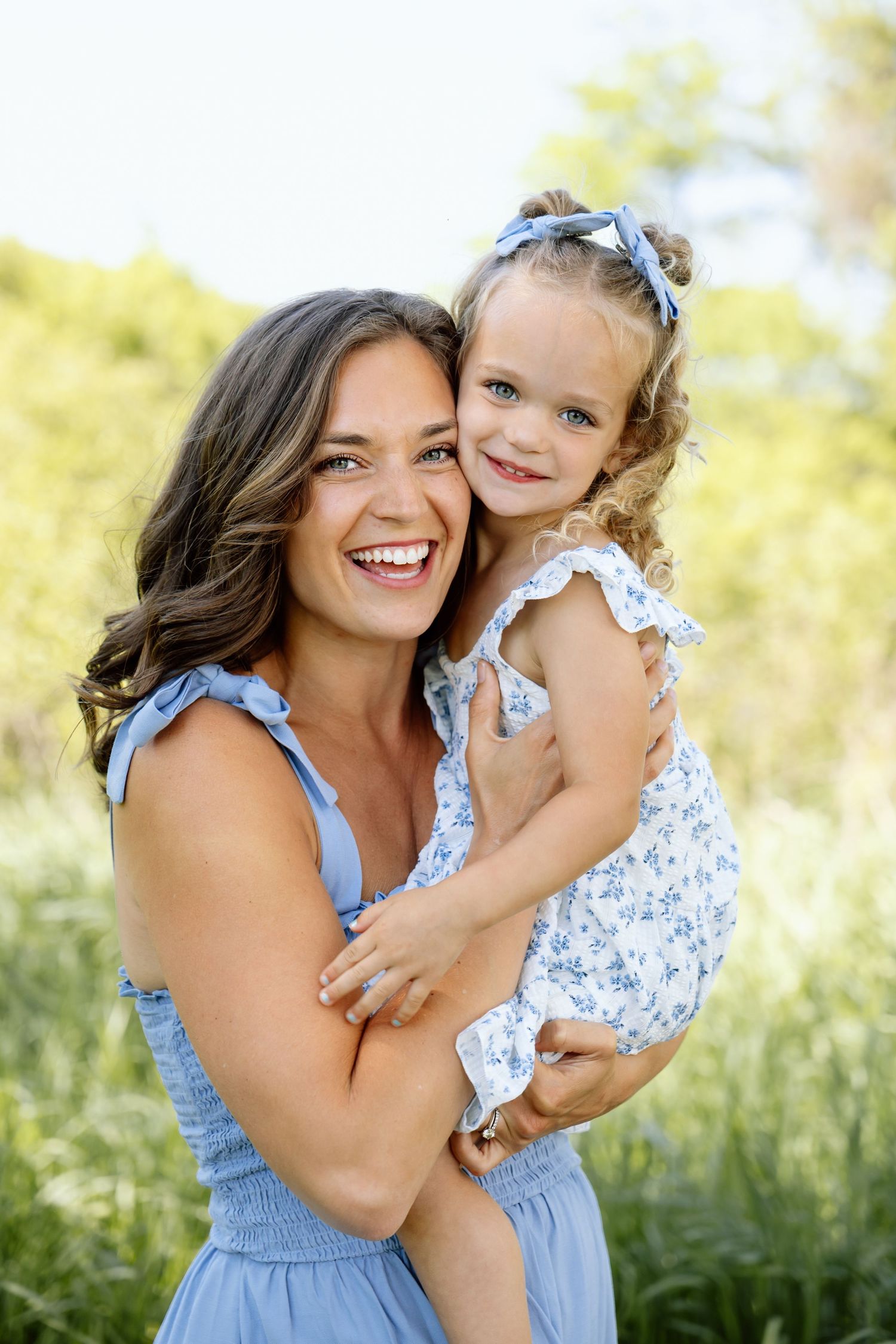 A smiling mother in a blue dress holds her young daughter outdoors in a sunny green field.