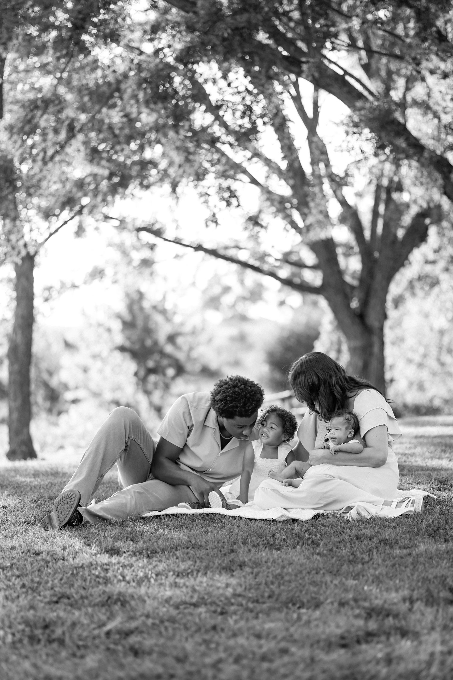 Black and white photo of family of four relaxing on blanket under trees.