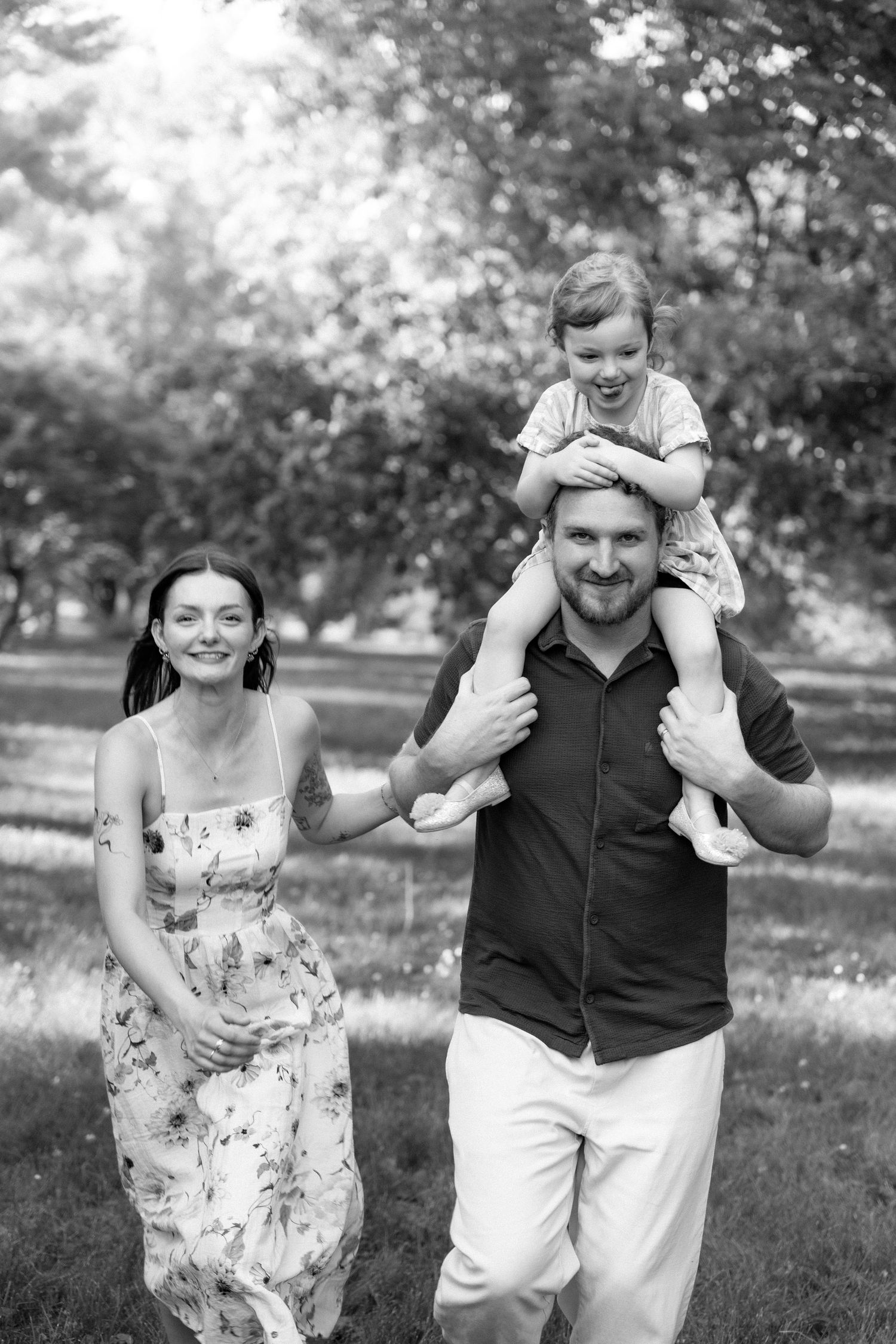 Black and white family portrait of parents walking with toddler on dad's shoulders in park.