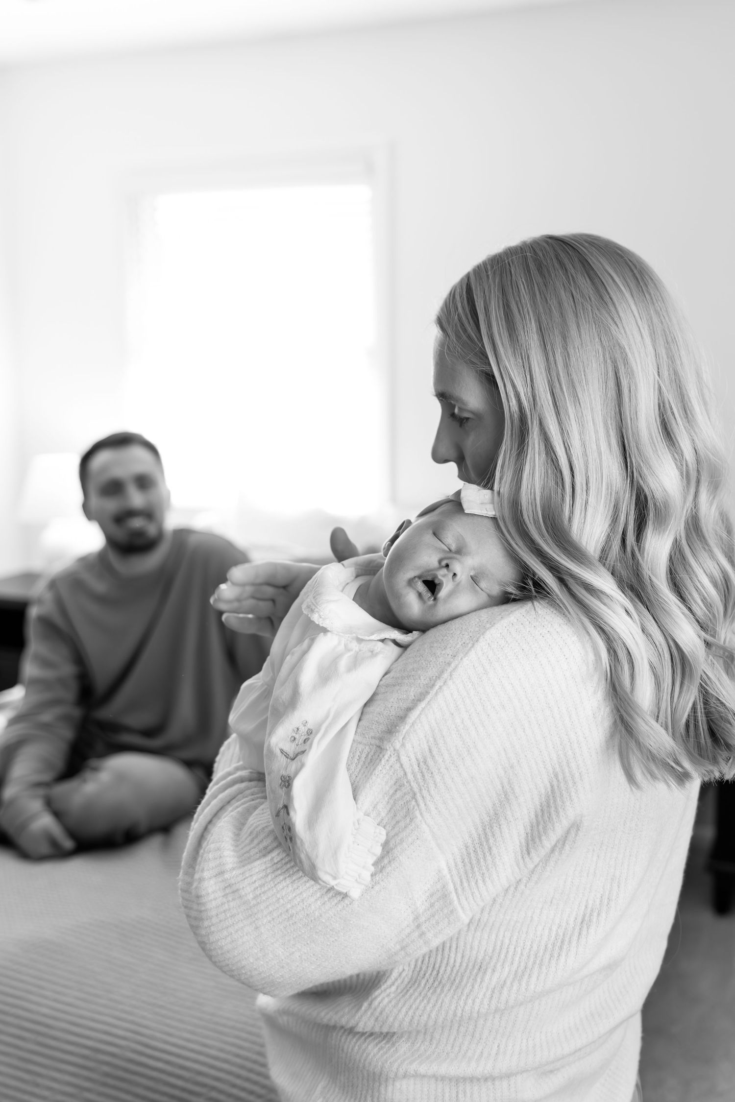 Black and white photo of a mother holding her newborn while father watches.