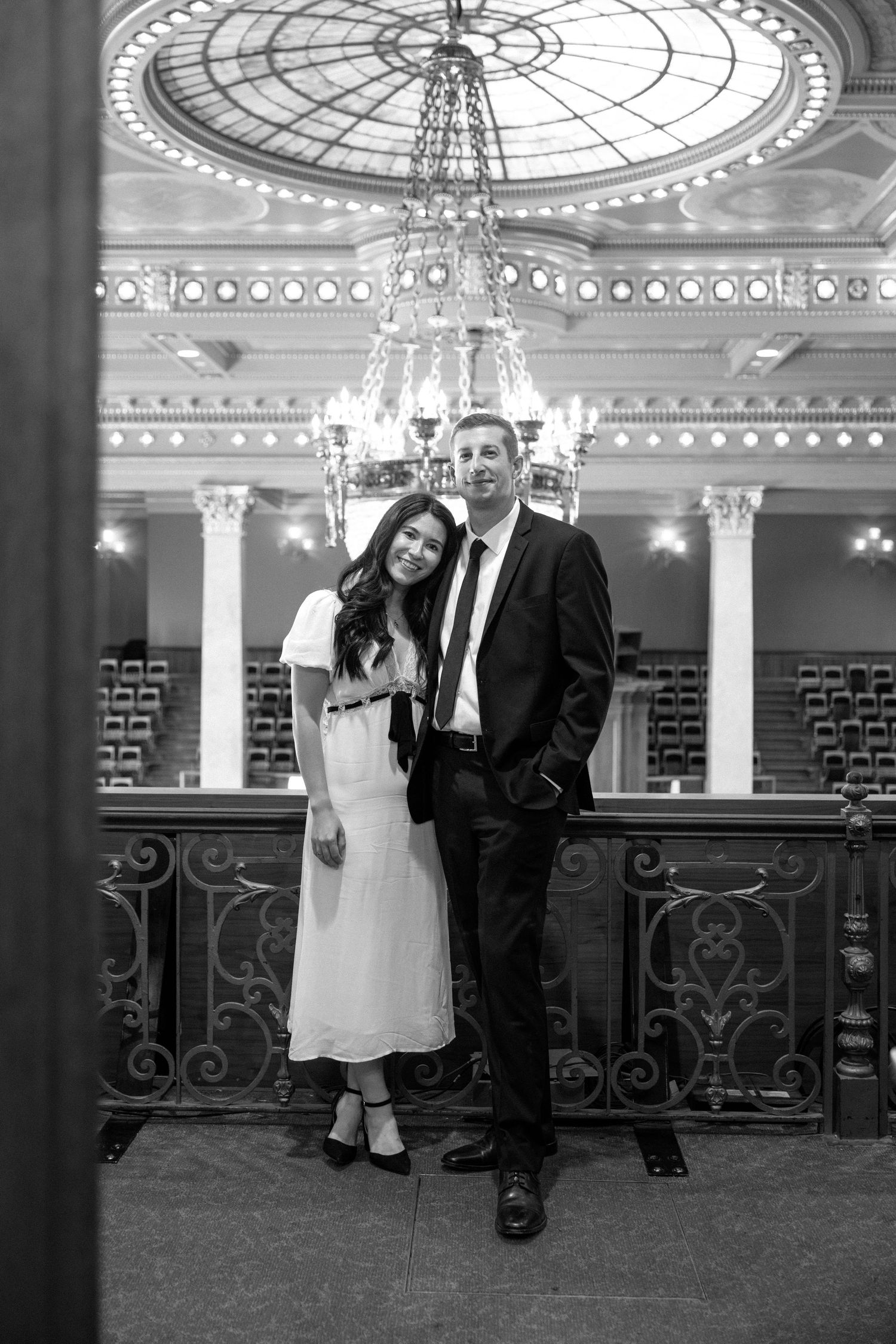 Black and white photo of couple posing beneath a stunning domed ceiling with crystal chandelier.