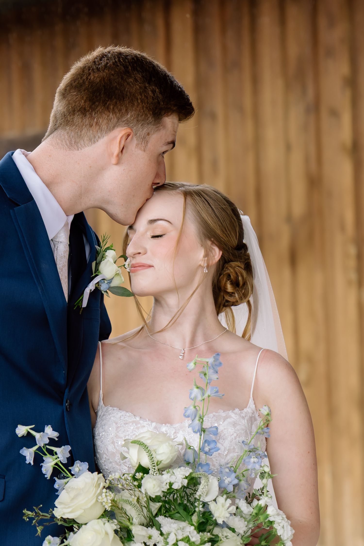 Groom in navy suit kissing bride on forehead as she holds white floral bouquet.