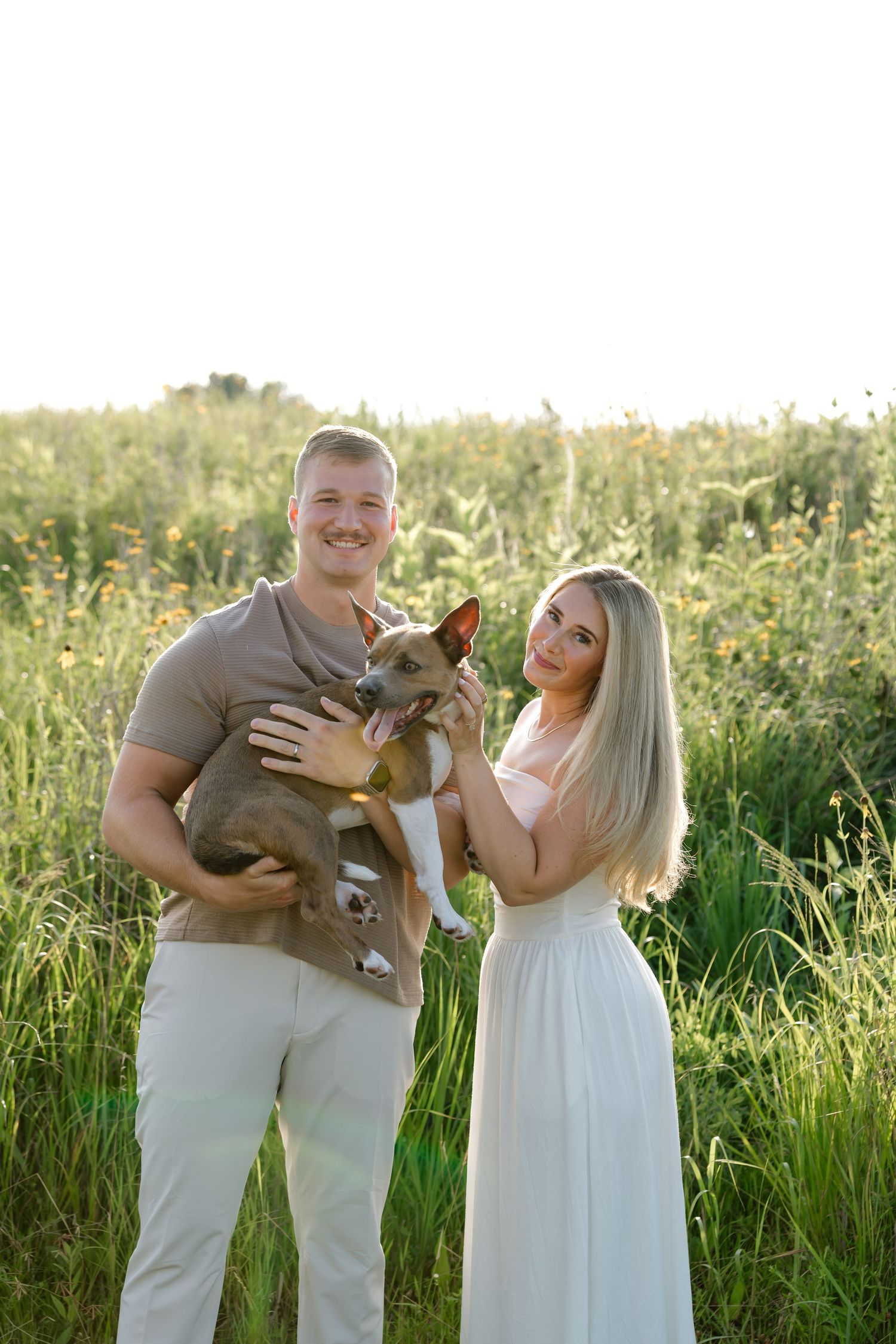 A smiling couple holds a brown dog in a sunlit green field, the woman in white and man in tan casual attire.