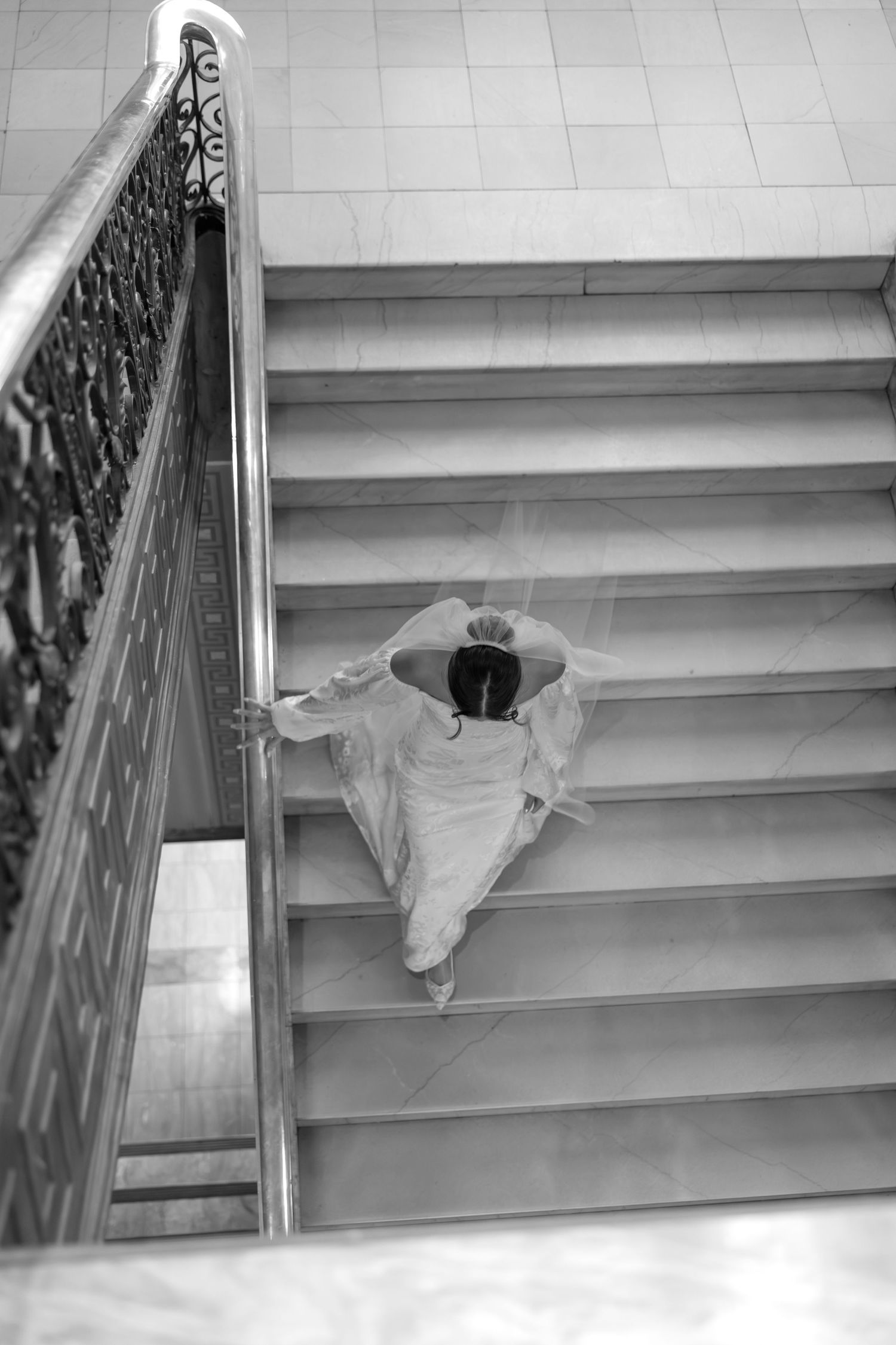Aerial black and white view of bride in white gown ascending marble staircase with iron railing.