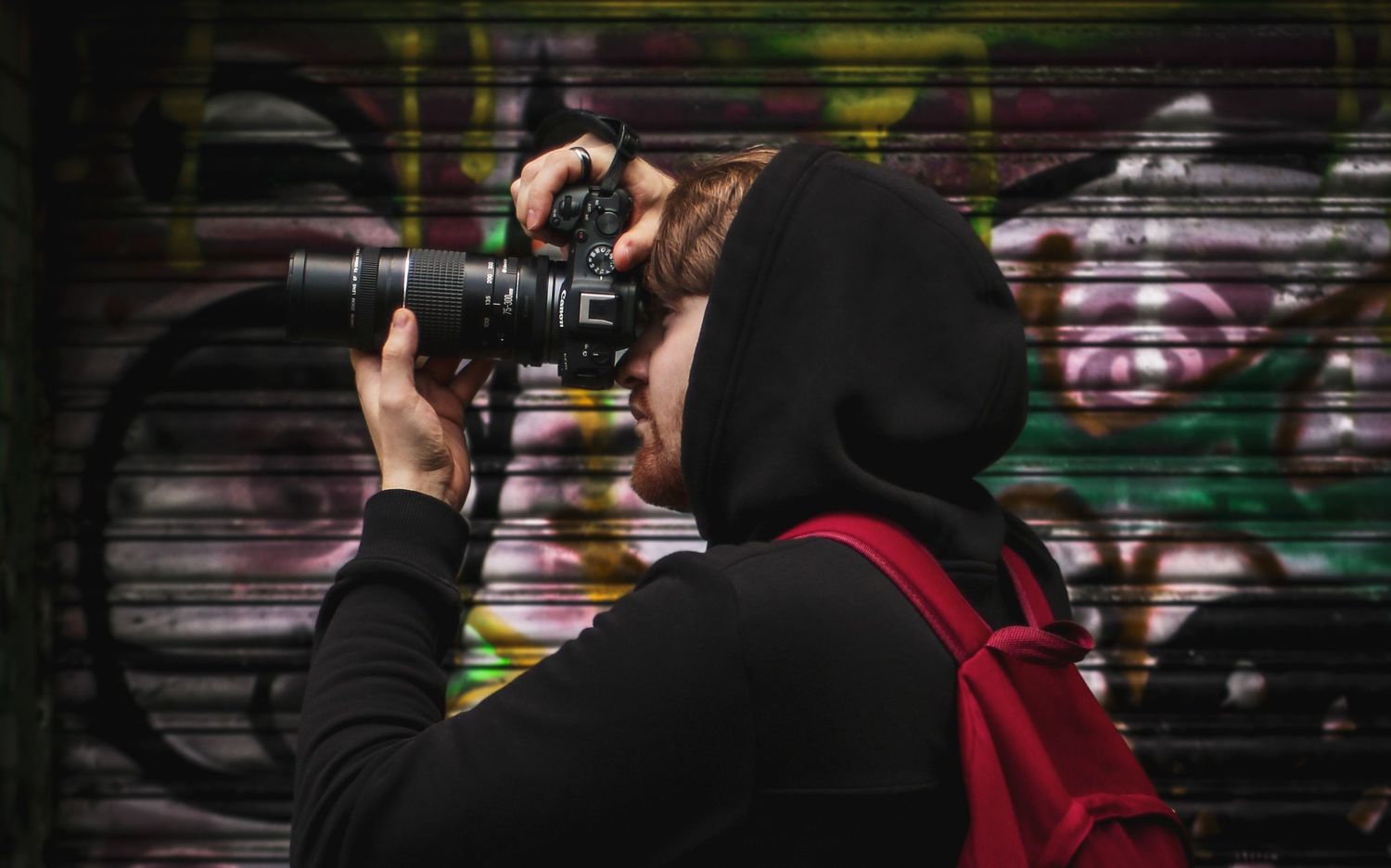 A hooded photographer captures images with a DSLR camera against a colorful graffiti wall backdrop.