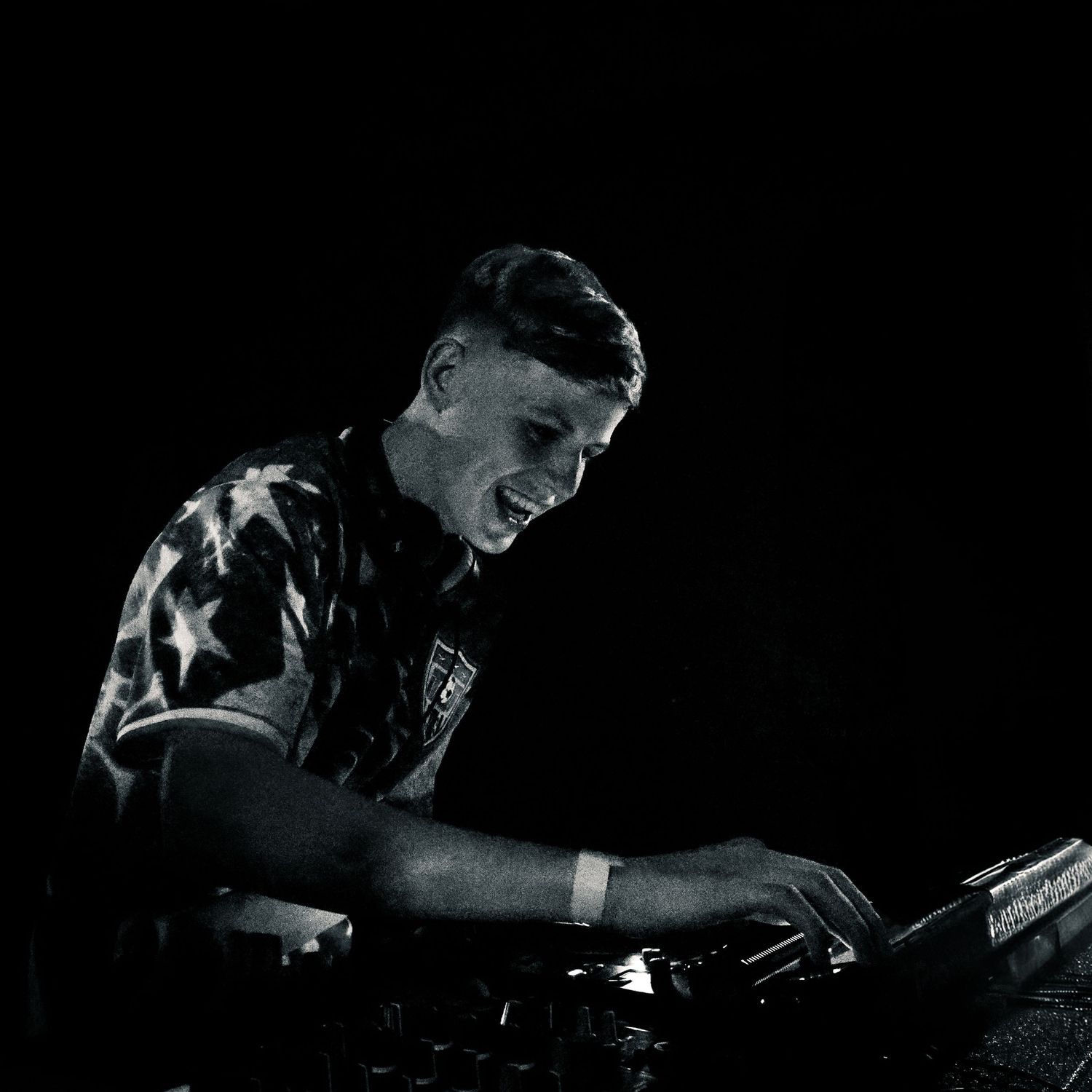 Black and white photo of a smiling DJ performing at turntables, wearing a star-patterned shirt in dark venue.