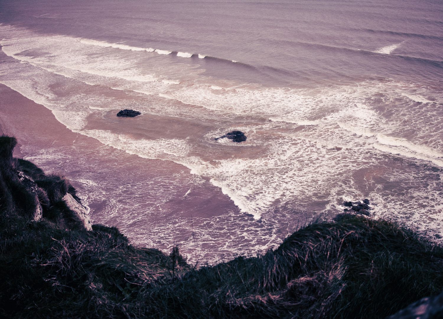 Aerial view of ocean waves crashing over dark rocks on a sandy shore.