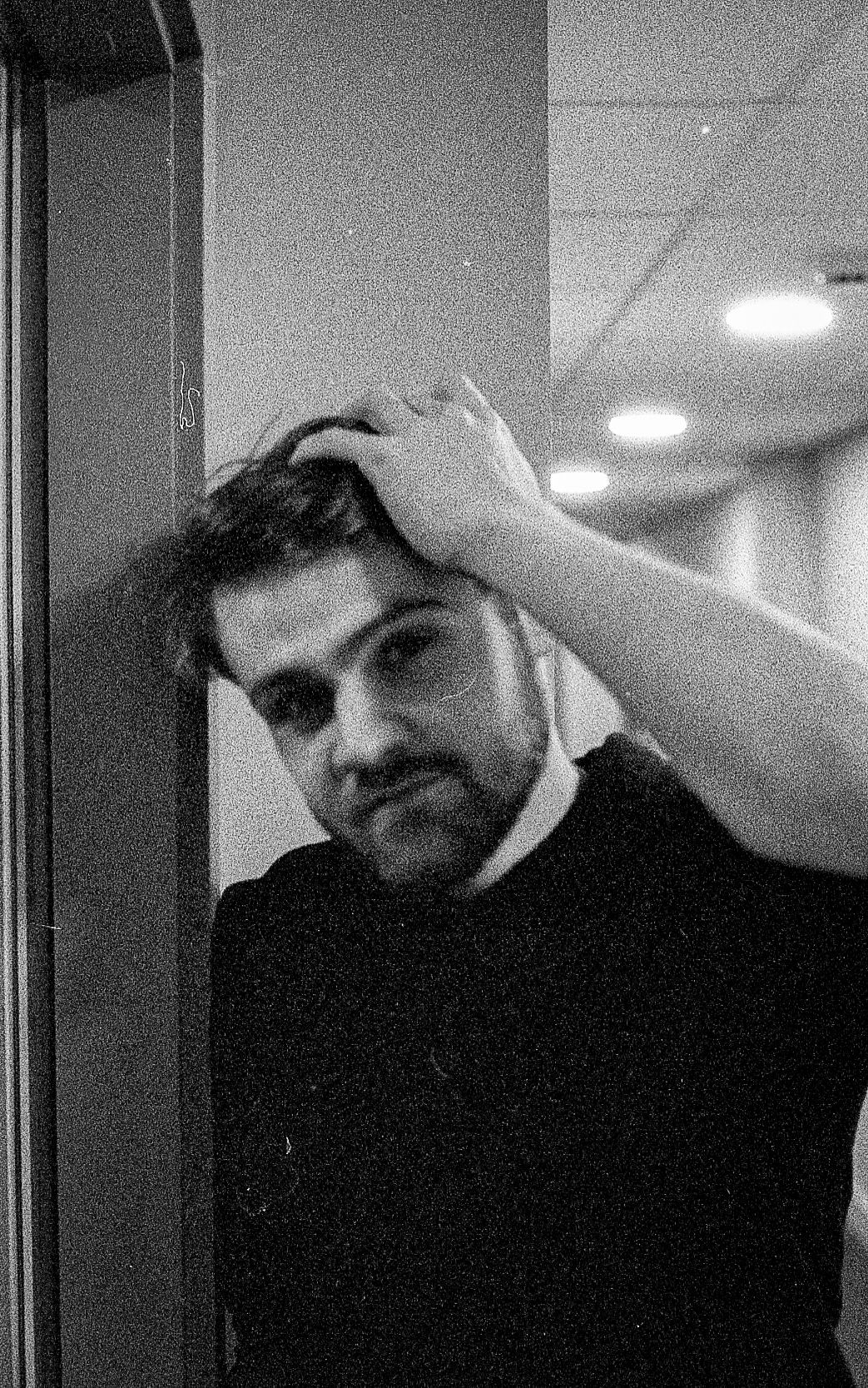 Black and white photo of a bearded man leaning against a wall, hand in hair, looking pensively at camera.