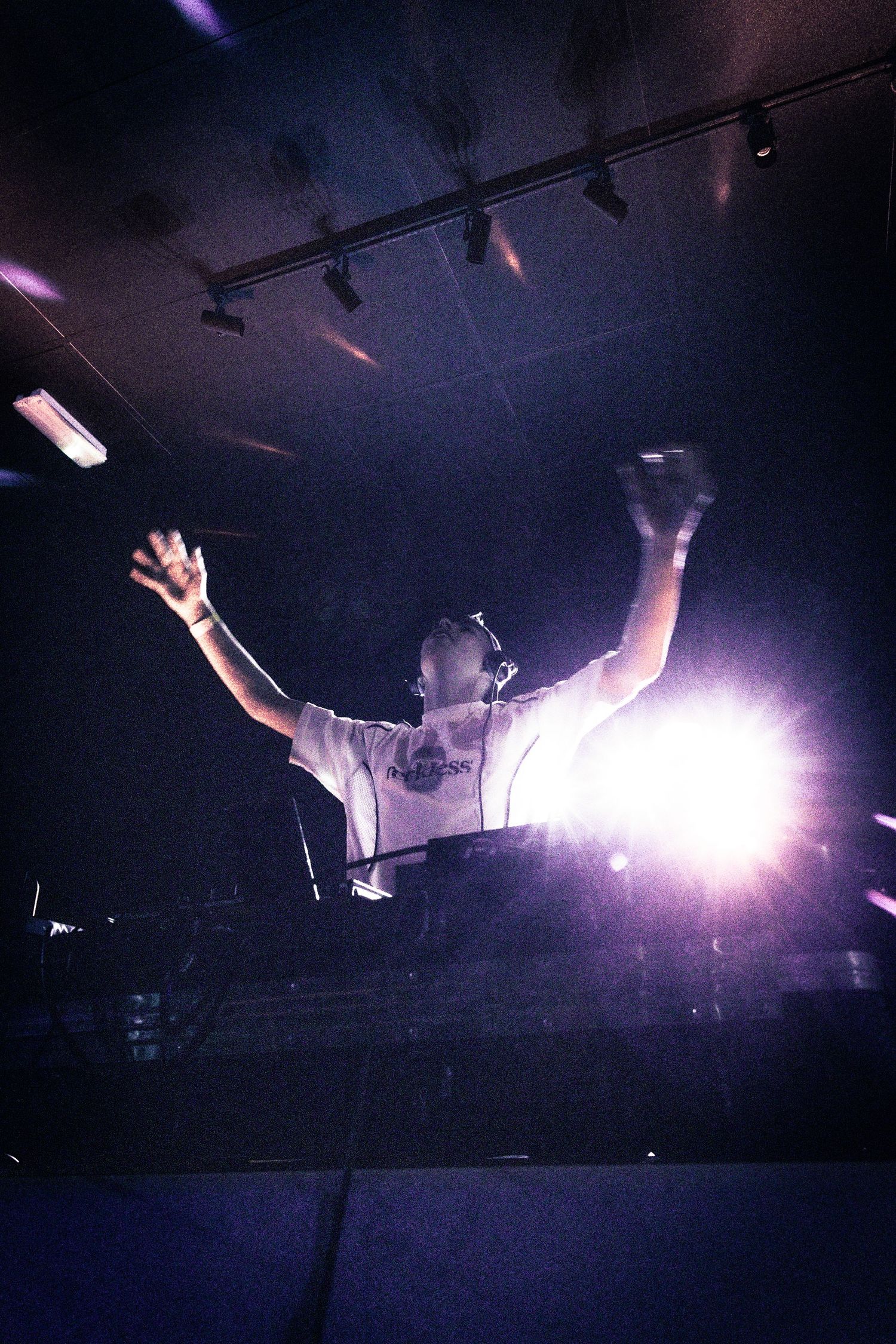 DJ performing with arms raised under bright stage lights in a dark club venue.