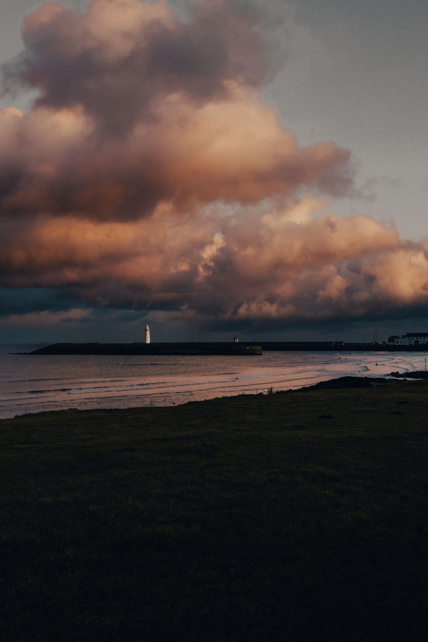 Dramatic sunset over coastal shoreline with a lighthouse silhouette beneath glowing orange storm clouds.