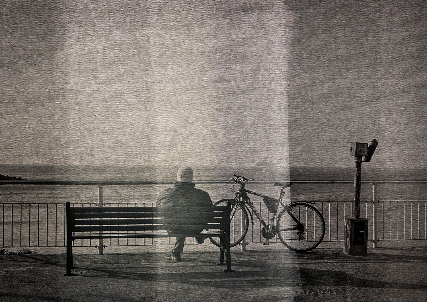 Person sitting alone on a bench by the seaside with a bicycle nearby, captured in moody black and white photography.