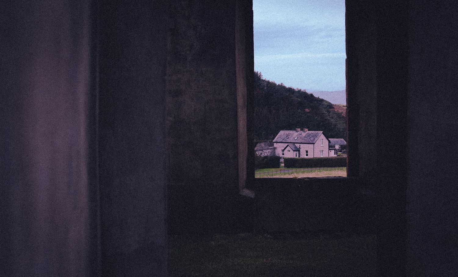 View through concrete ruins framing a pink farmhouse against a hillside at dusk.
