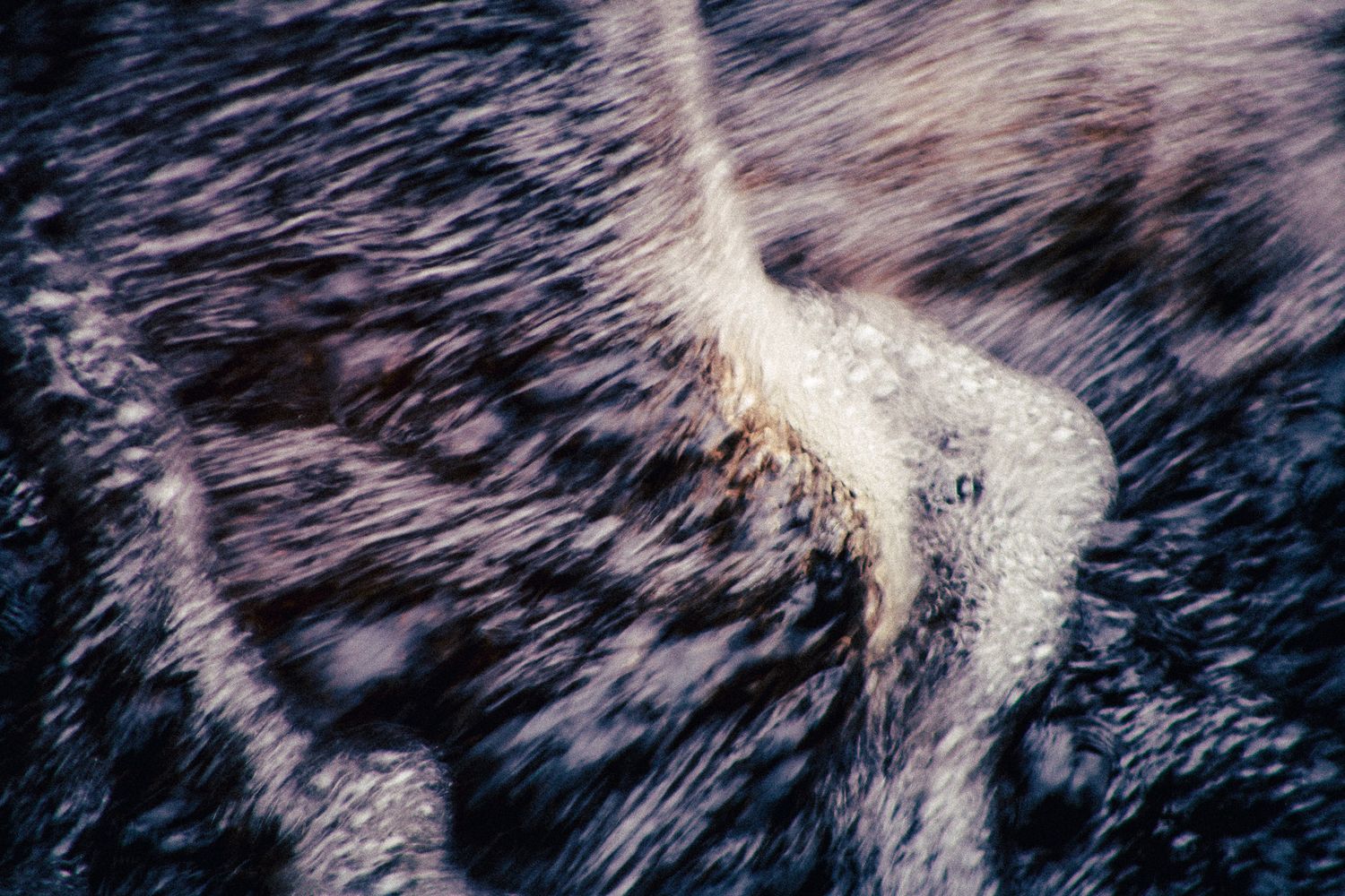 Close-up aerial view of rushing water flowing over rocks, creating swirling white foam patterns in a natural stream.