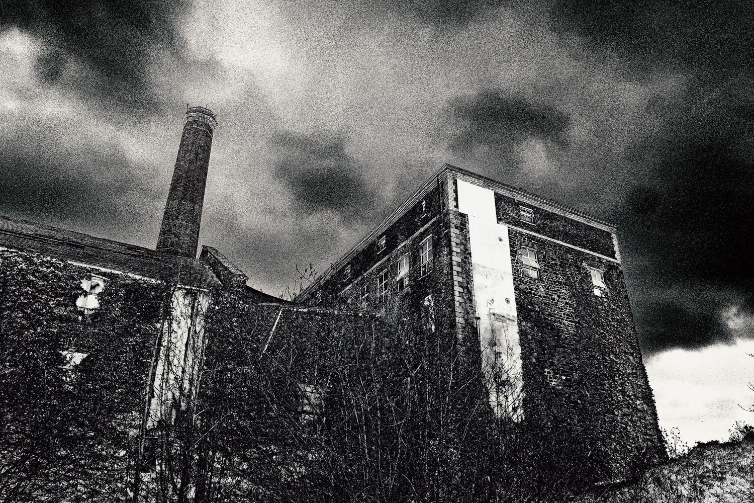 Dramatic black and white photo of an abandoned industrial building with ivy-covered walls under stormy skies.