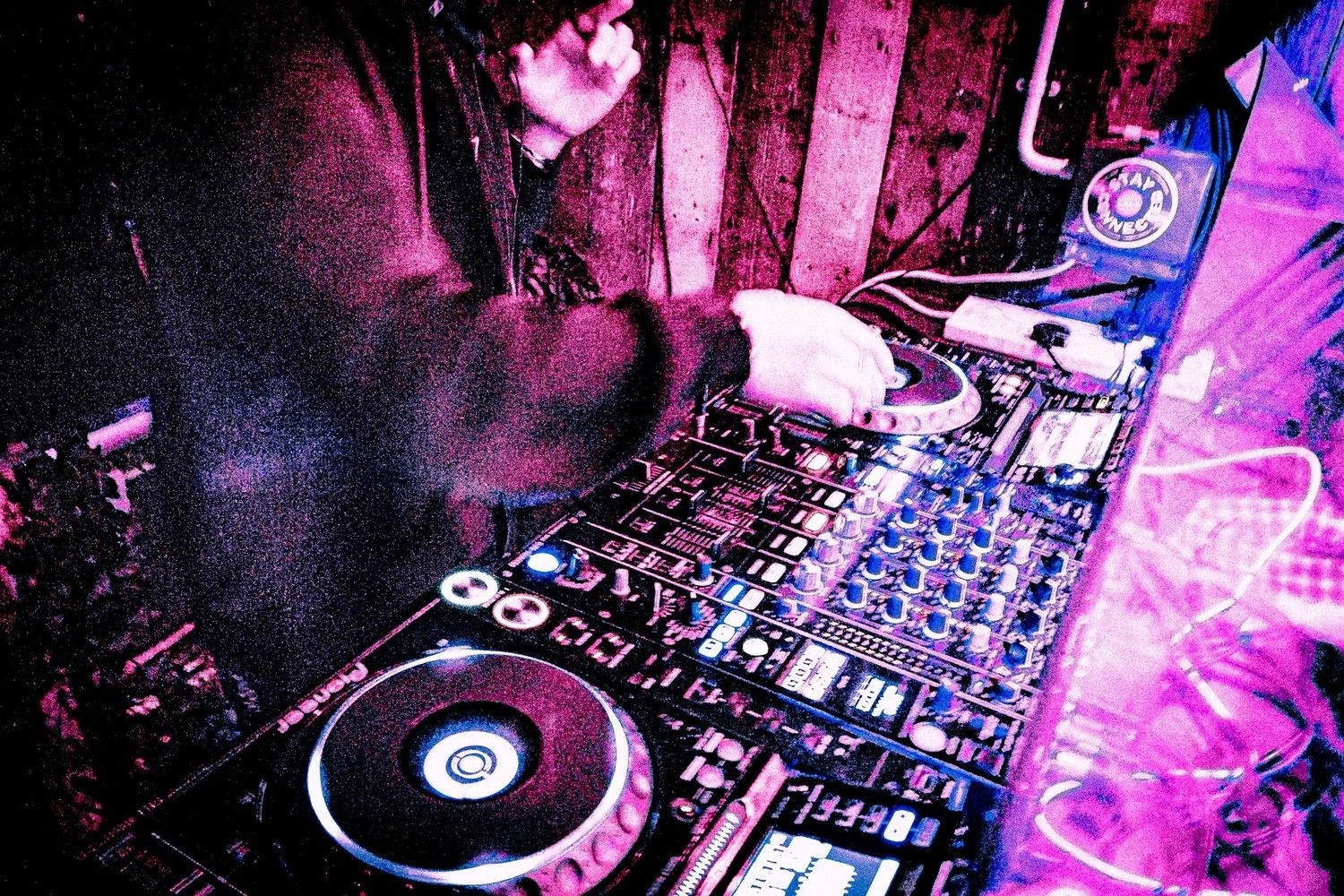 DJ performing at a nightclub with Pioneer CDJ decks under vibrant pink and purple stage lighting.