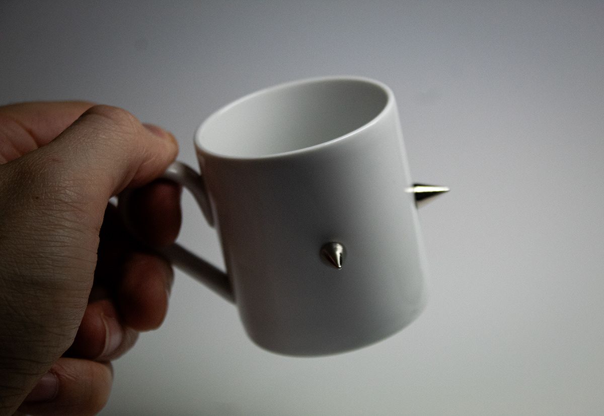 White ceramic mug with silver metal spikes held by a hand against a gray background.