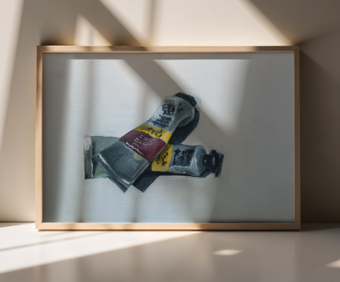 Framed artwork of paint tubes in a wooden frame, displayed against a sunlit wall with soft shadows.