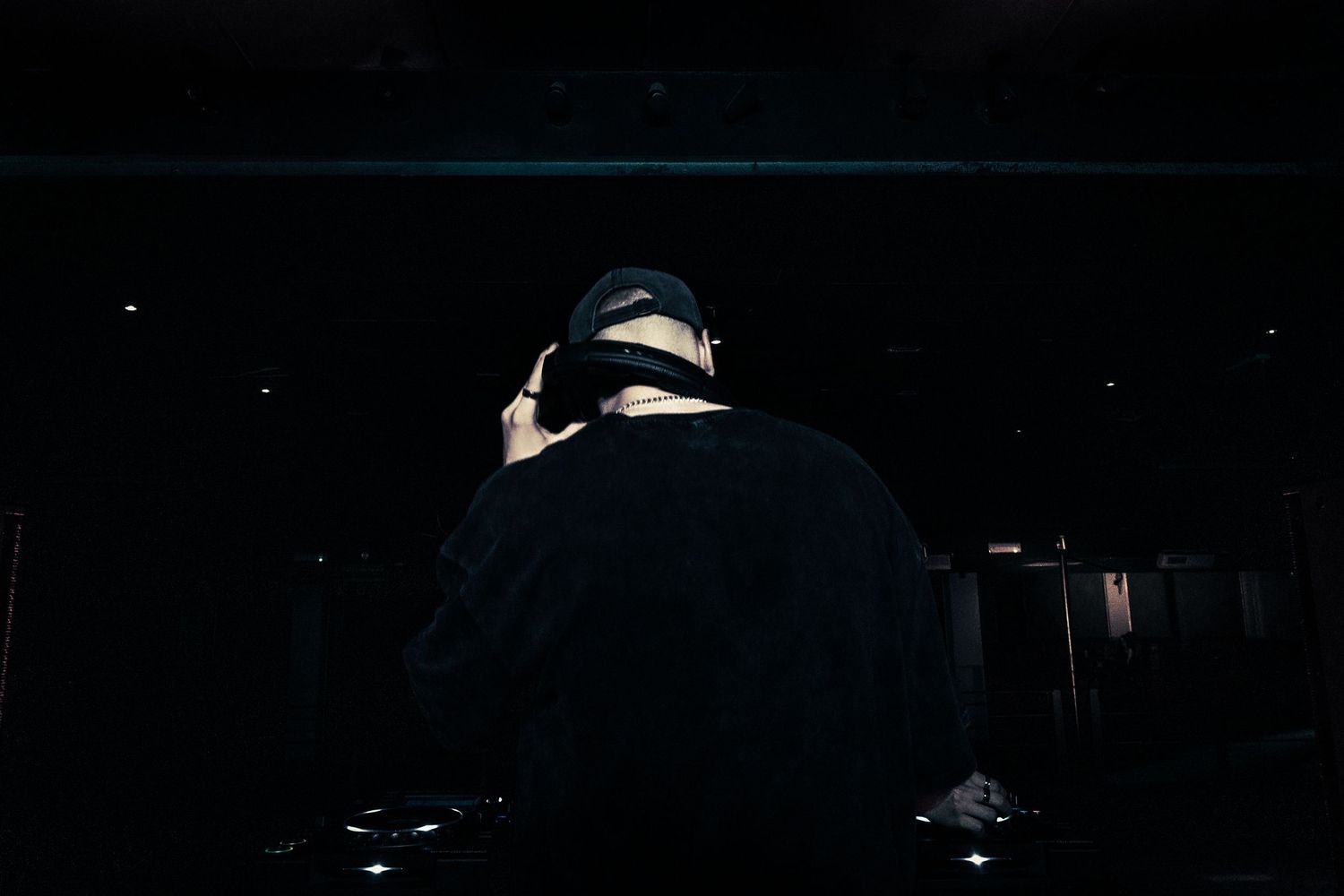 DJ performing in dark club, wearing headphones and cap, silhouetted against dimly lit stage equipment.