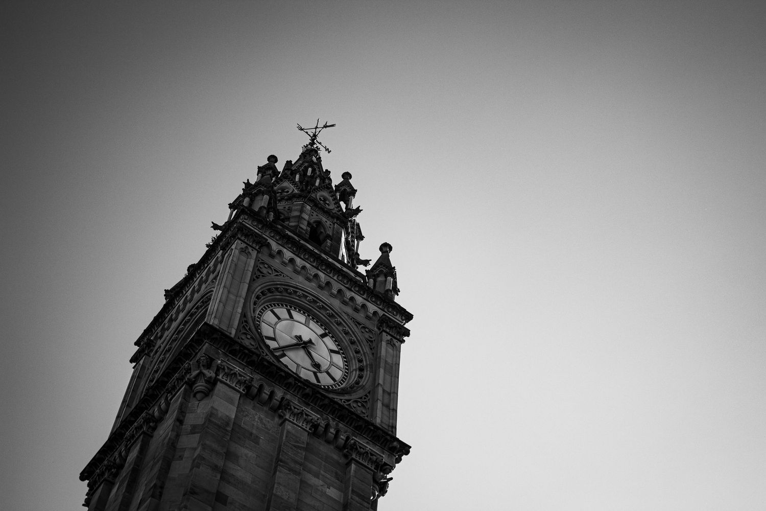 Black and white photo of a historic Victorian clock tower with ornate stone carvings and a cross at the top.