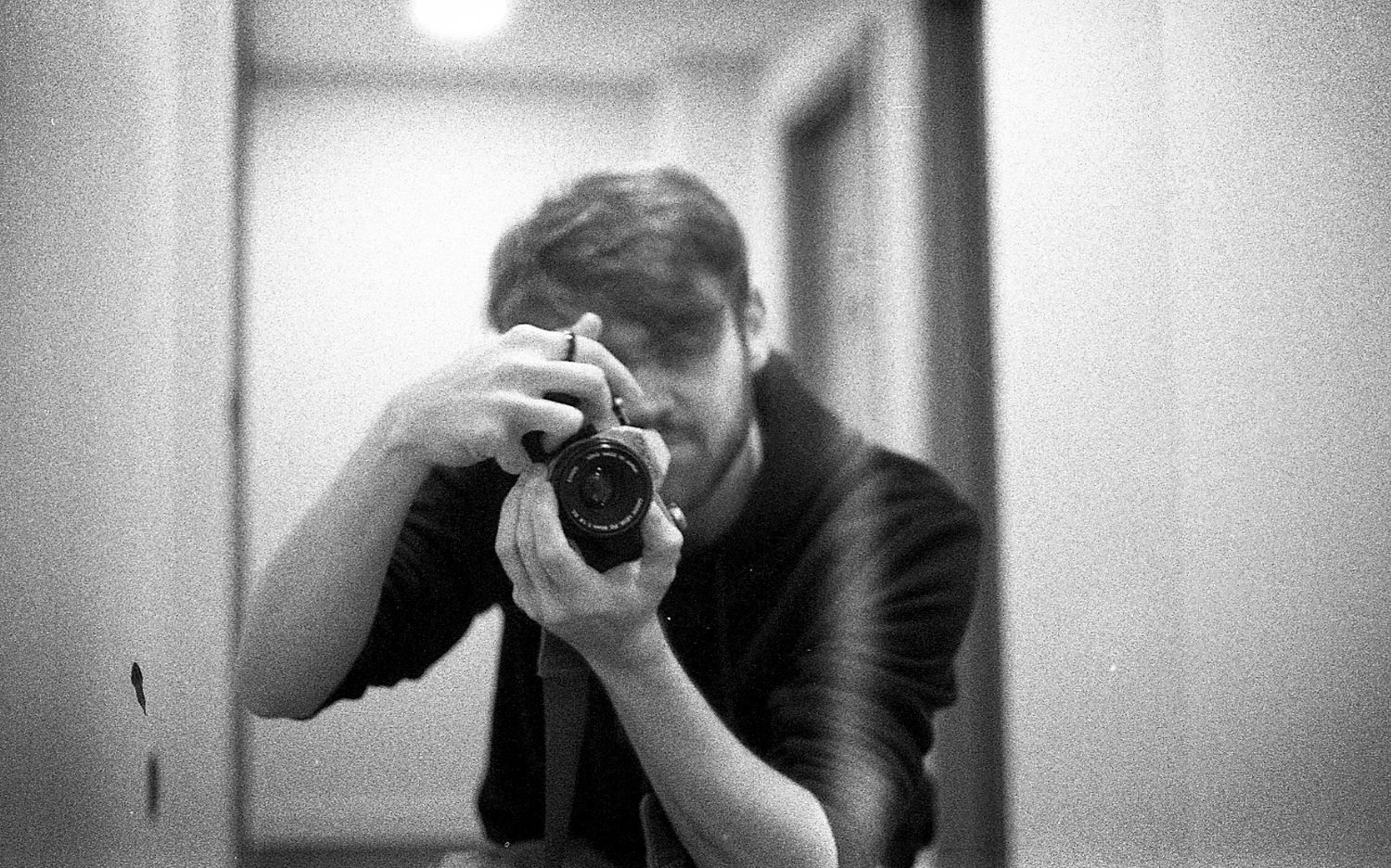 Black and white self-portrait of a man taking a photo in a mirror with a camera covering his face.