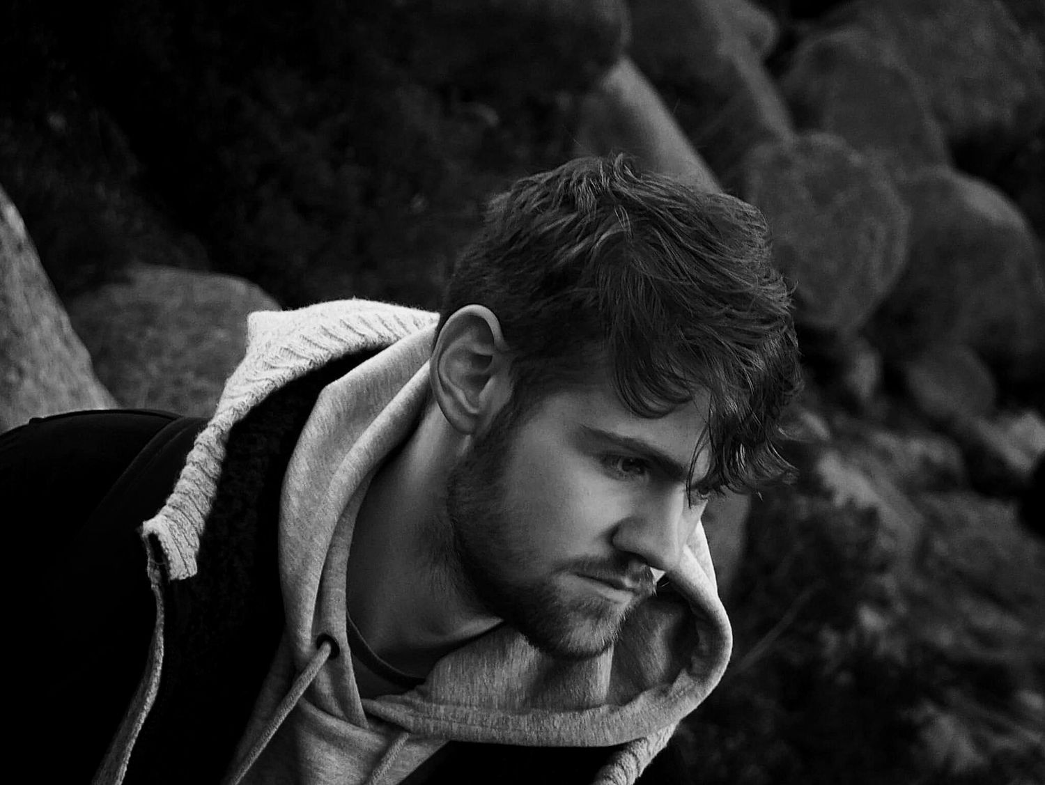 Black and white portrait of a young man in a hoodie leaning forward against rocky background.