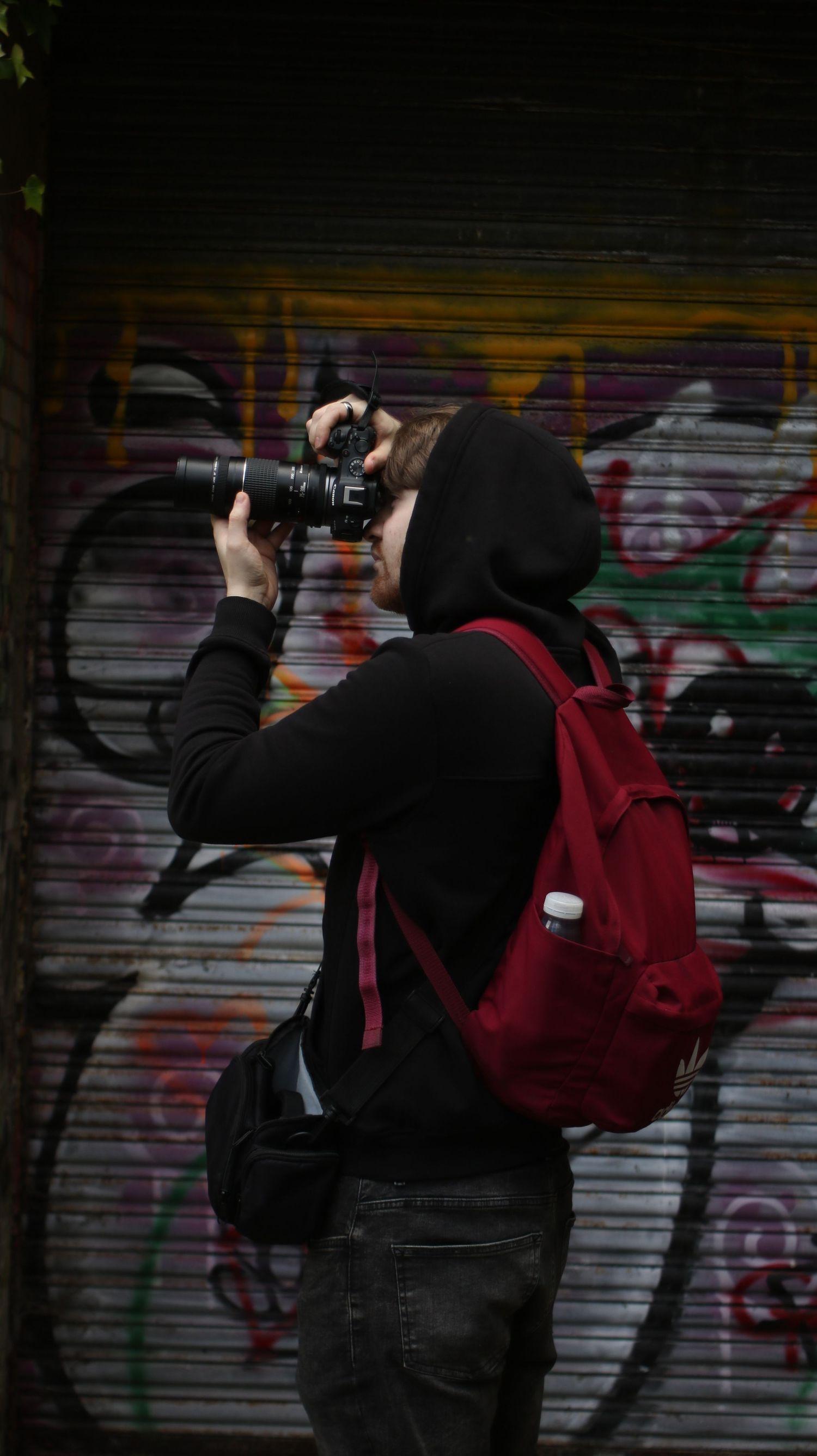 Person in black hoodie with red backpack photographing graffiti-covered metal shutters with a DSLR camera.
