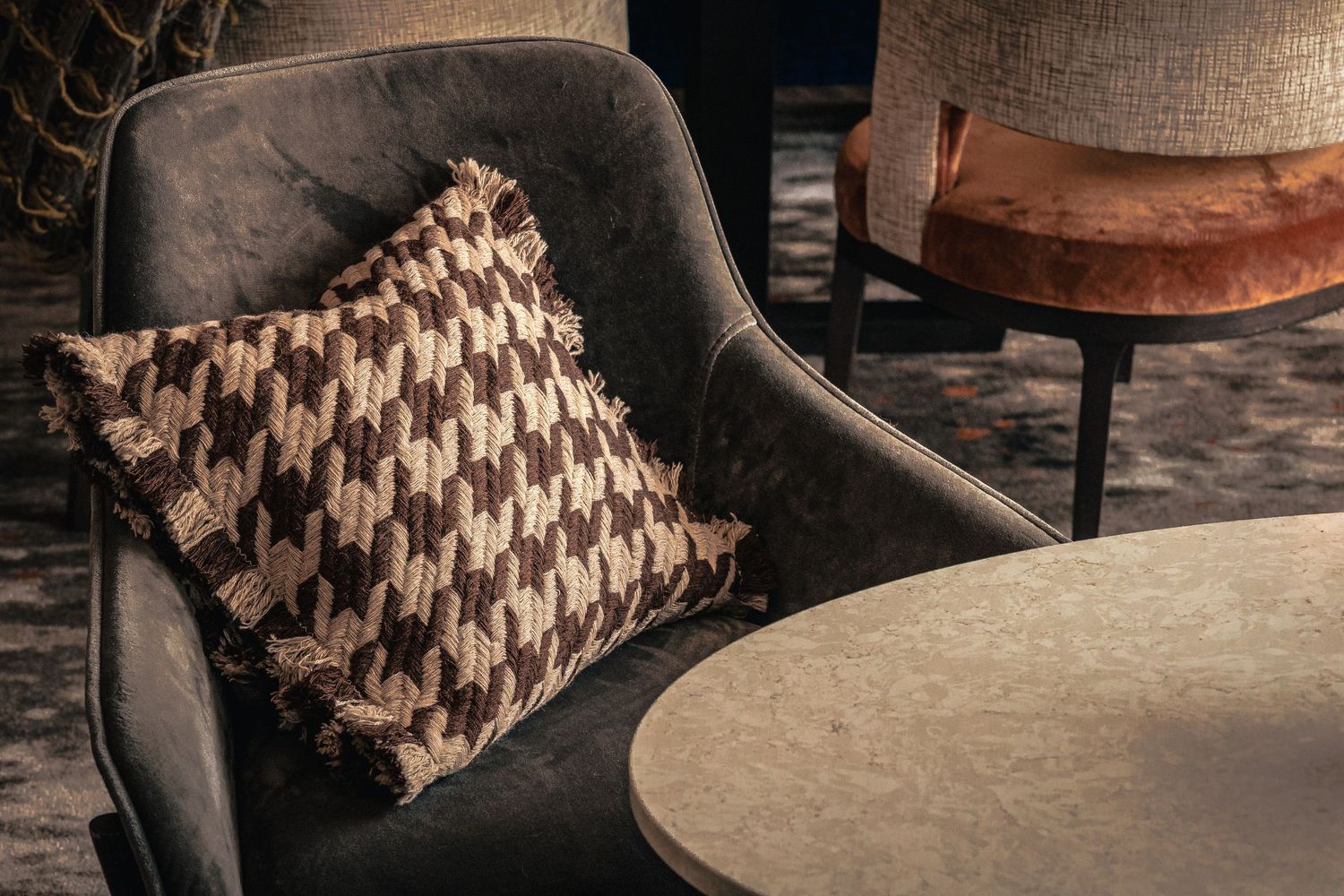Dark velvet armchair with houndstooth fringe pillow beside marble coffee table in elegant interior setting.